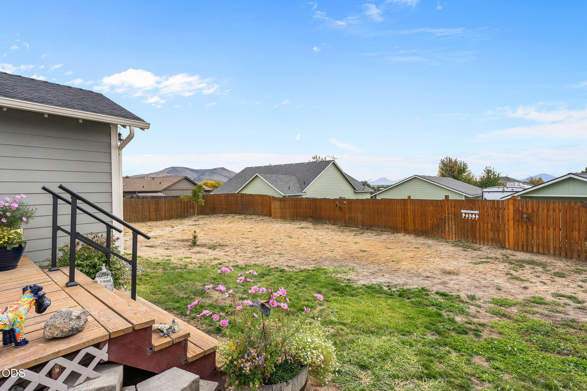 Fenced backyard with a dry lawn, pink flowers, wooden deck, and distant hills.