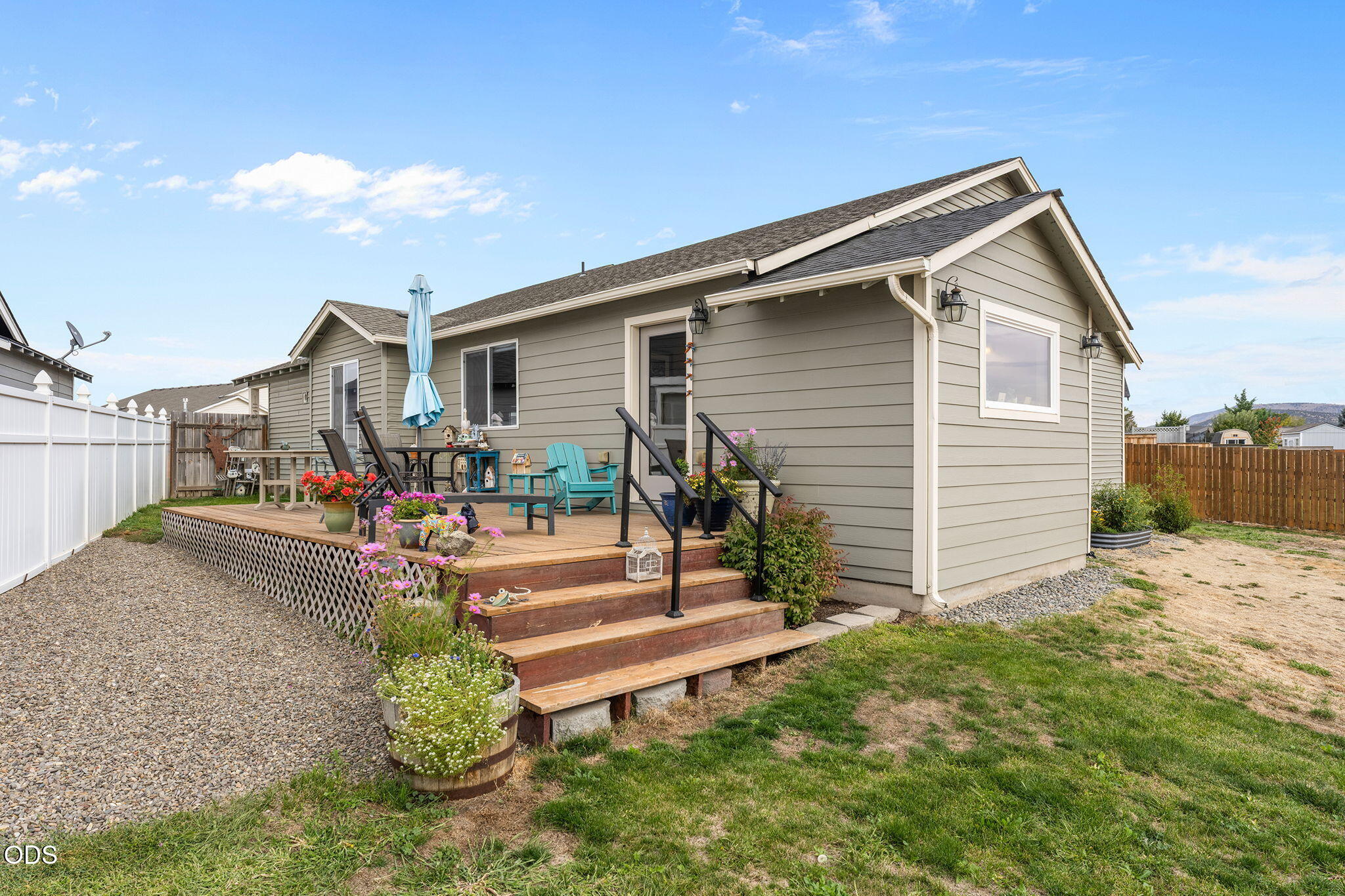 Home’s rear deck with wooden stairs, patio seating, potted flowers, and a fenced backyard.