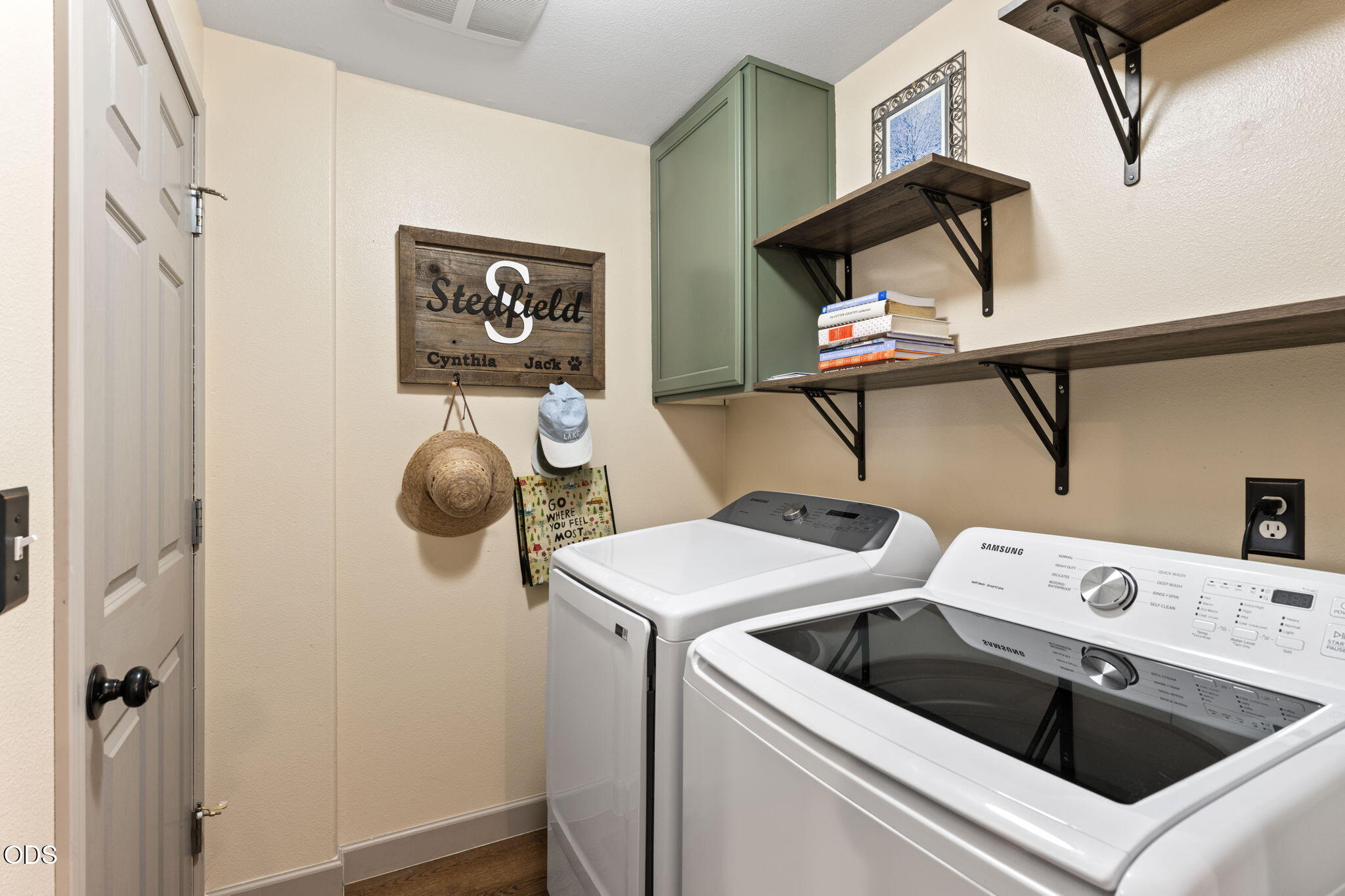 Laundry room with a white washer and Samsung dryer beneath wood shelves and a sage-green cabinet.