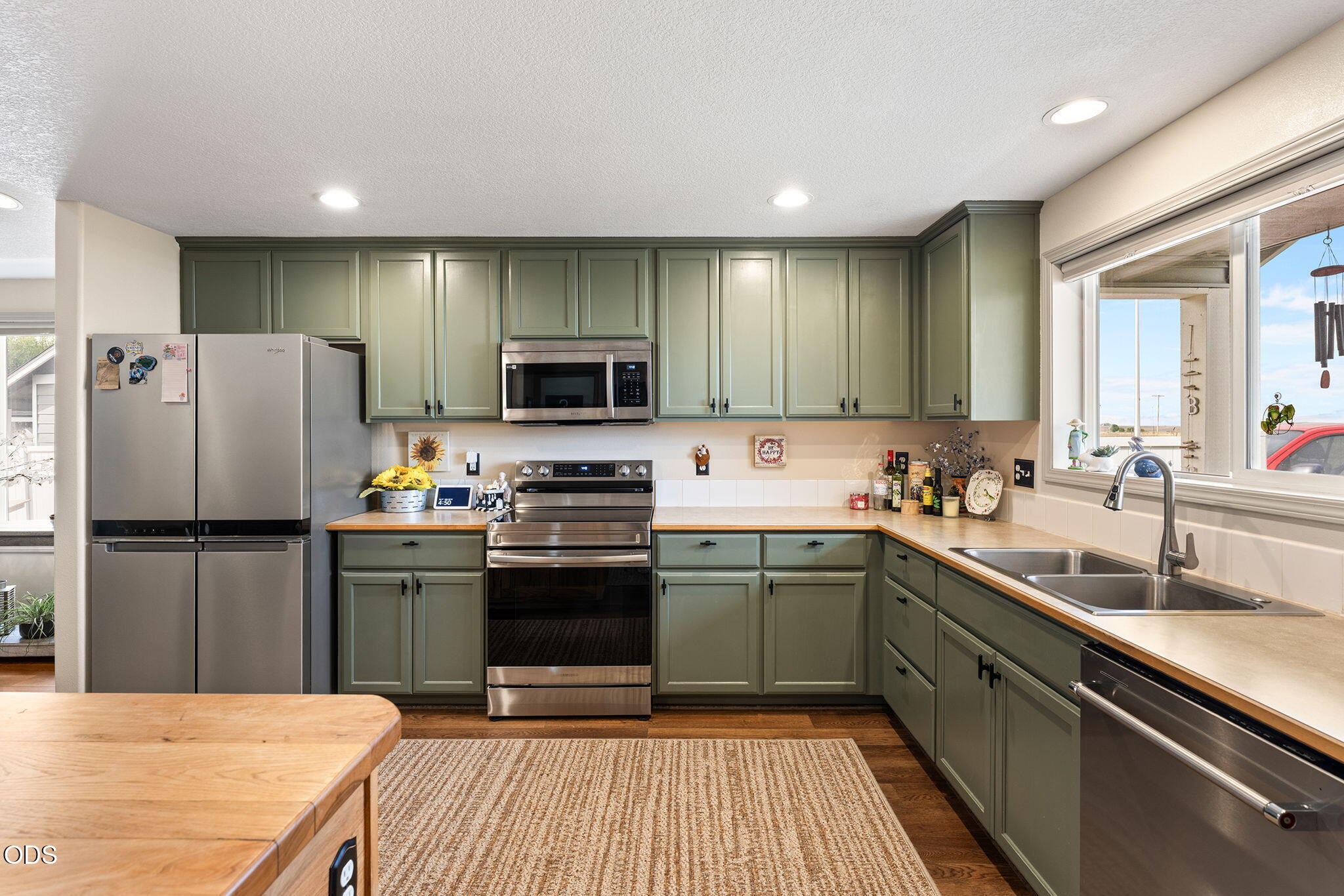 Kitchen with sage-green cabinets, stainless steel appliances, and a double-basin sink beneath a large window.
