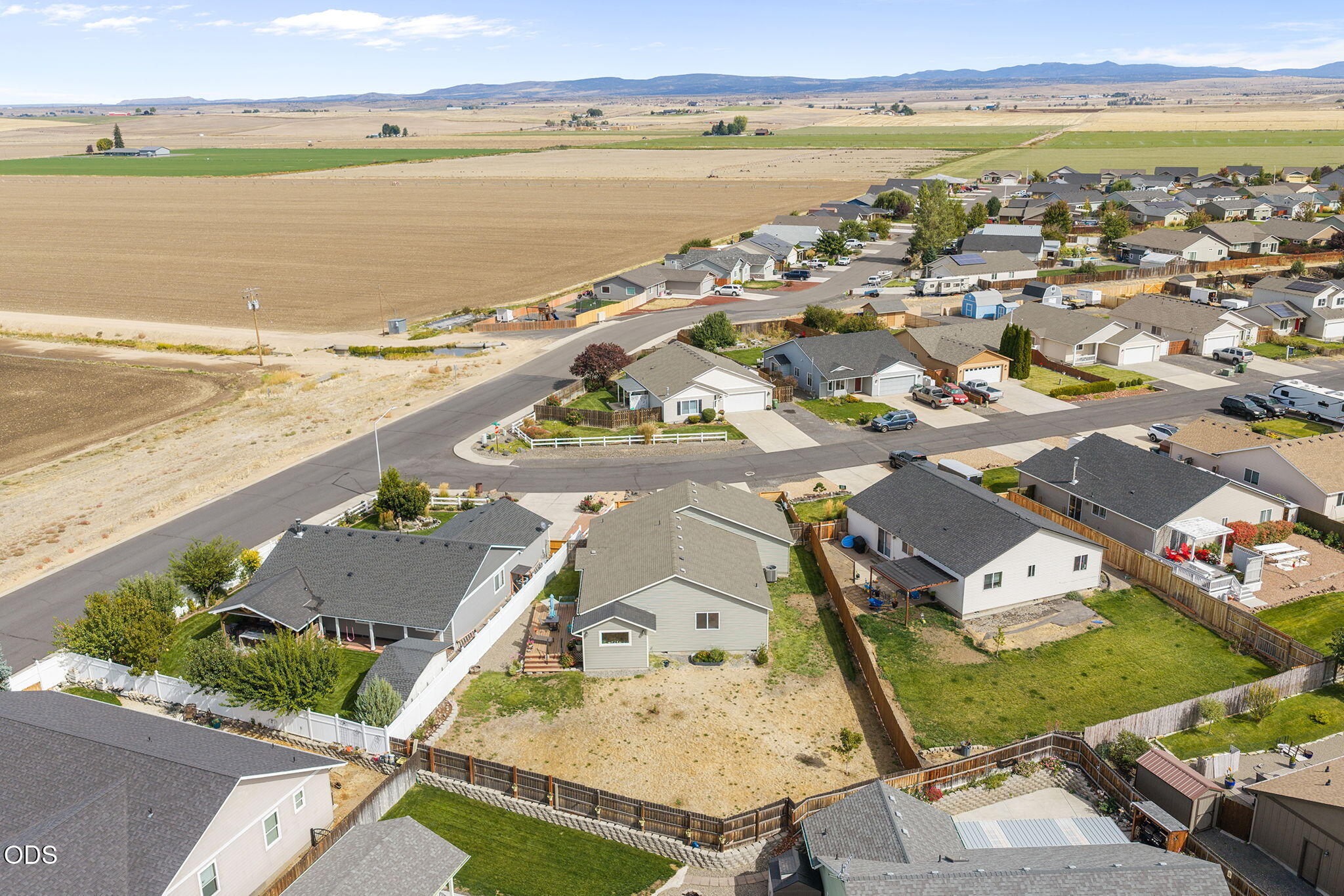 Aerial neighborhood view with fenced yards, agricultural fields, and distant mountains.