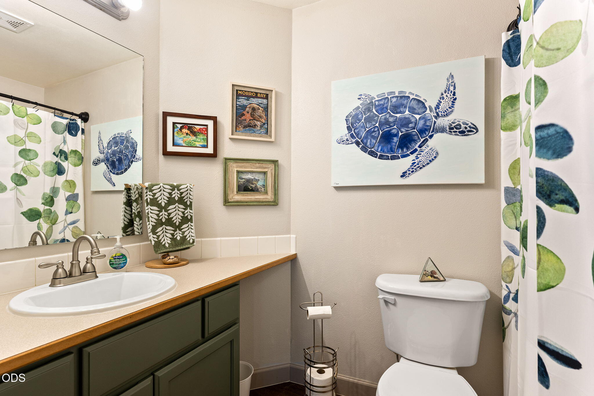 Bathroom with a dark green vanity, leafy shower curtain, and large blue sea turtle artwork.
