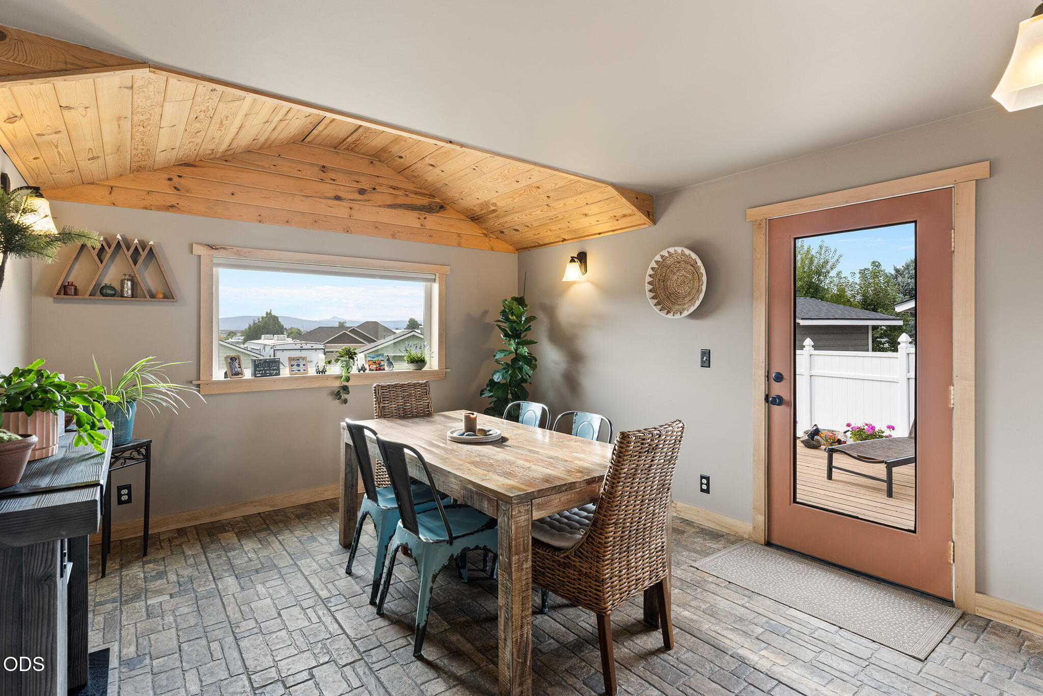 Dining room with a peaked wood-plank ceiling, rustic table, and glass door to a fenced deck.