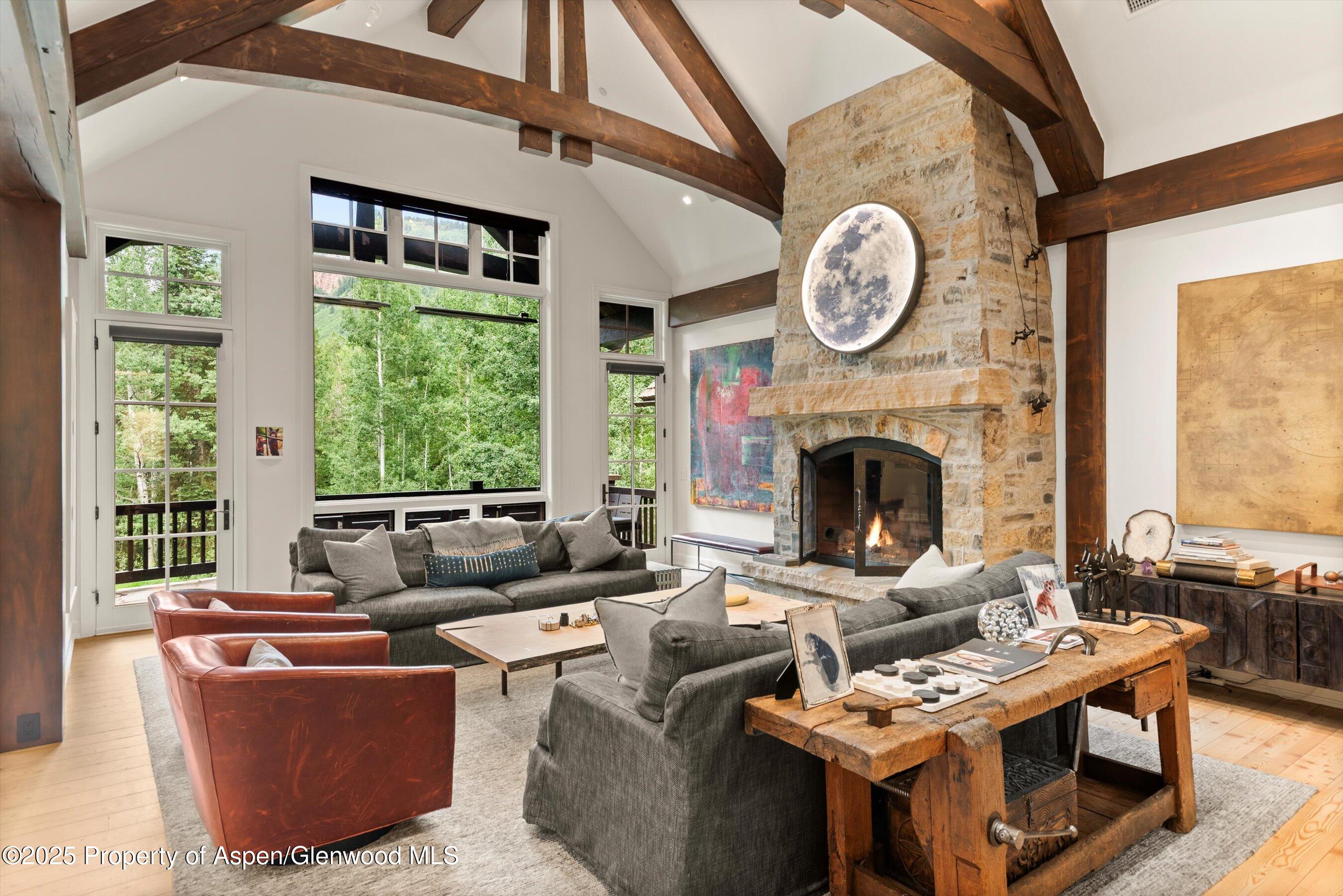 Living room with exposed timber beams, a stone fireplace, and large windows overlooking trees.