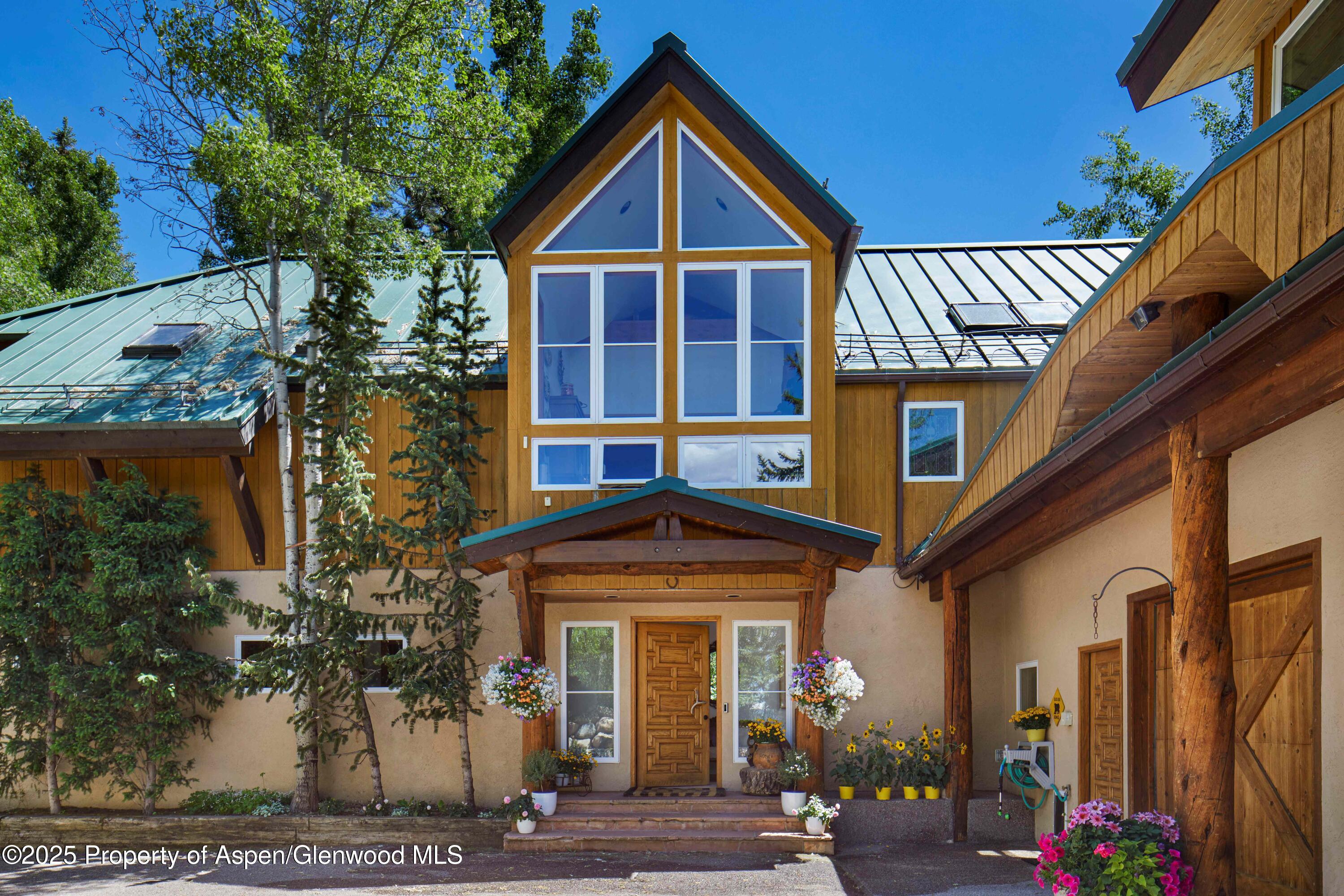 Wood-and-stucco home with tall gabled windows, green metal roof, and flower-lined entrance.