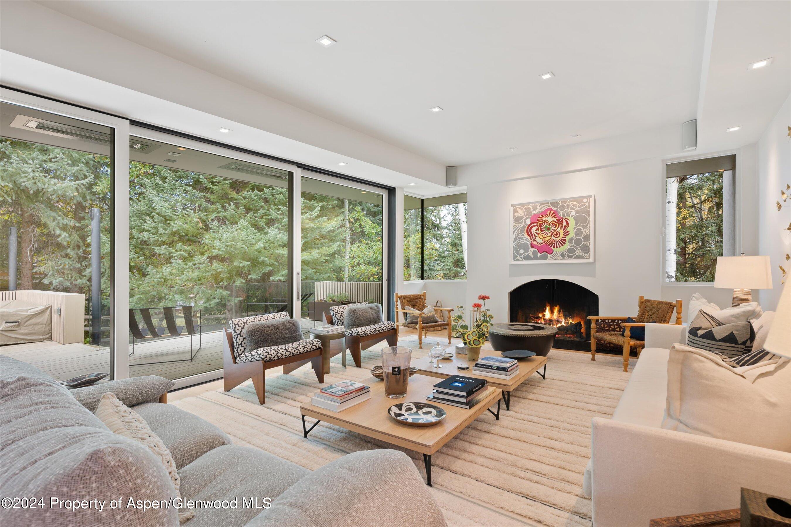 Living room with a lit fireplace and floor-to-ceiling glass doors overlooking a wooded deck.
