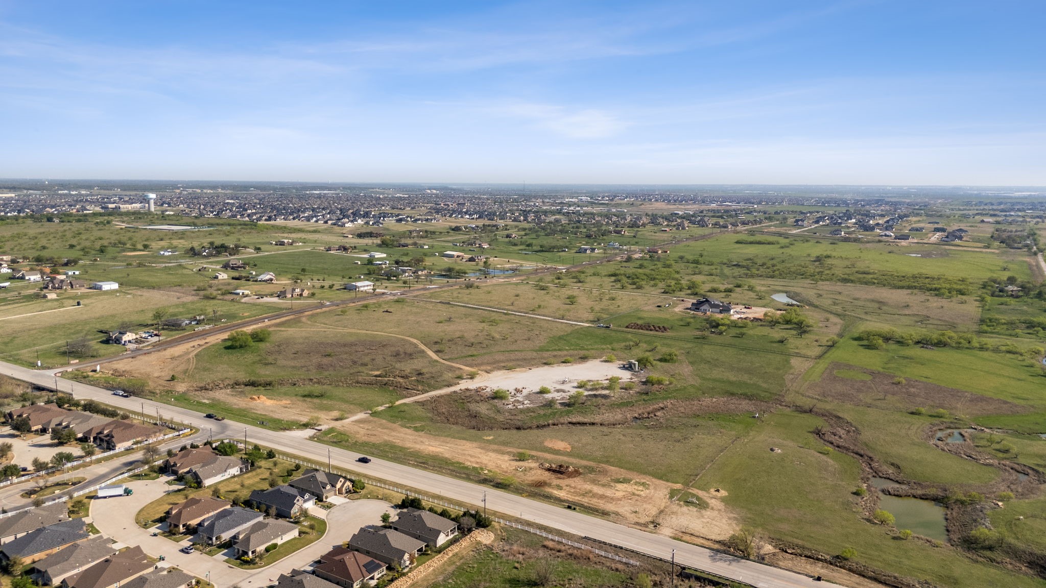 Aerial view of open land and scattered homes, with a suburban neighborhood on the horizon.