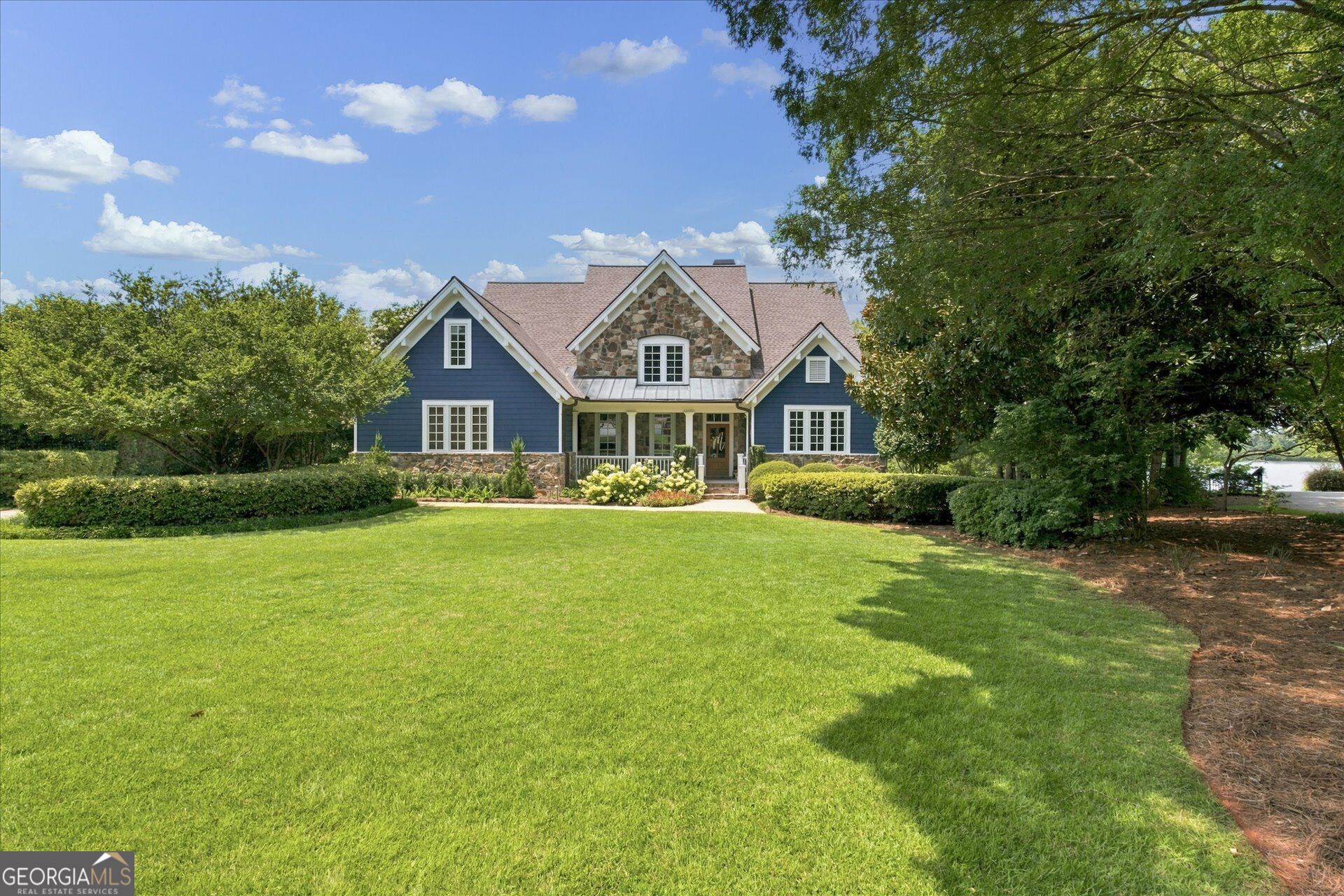 Blue-sided house with stone accents and a covered porch, framed by mature trees and a wide lawn.