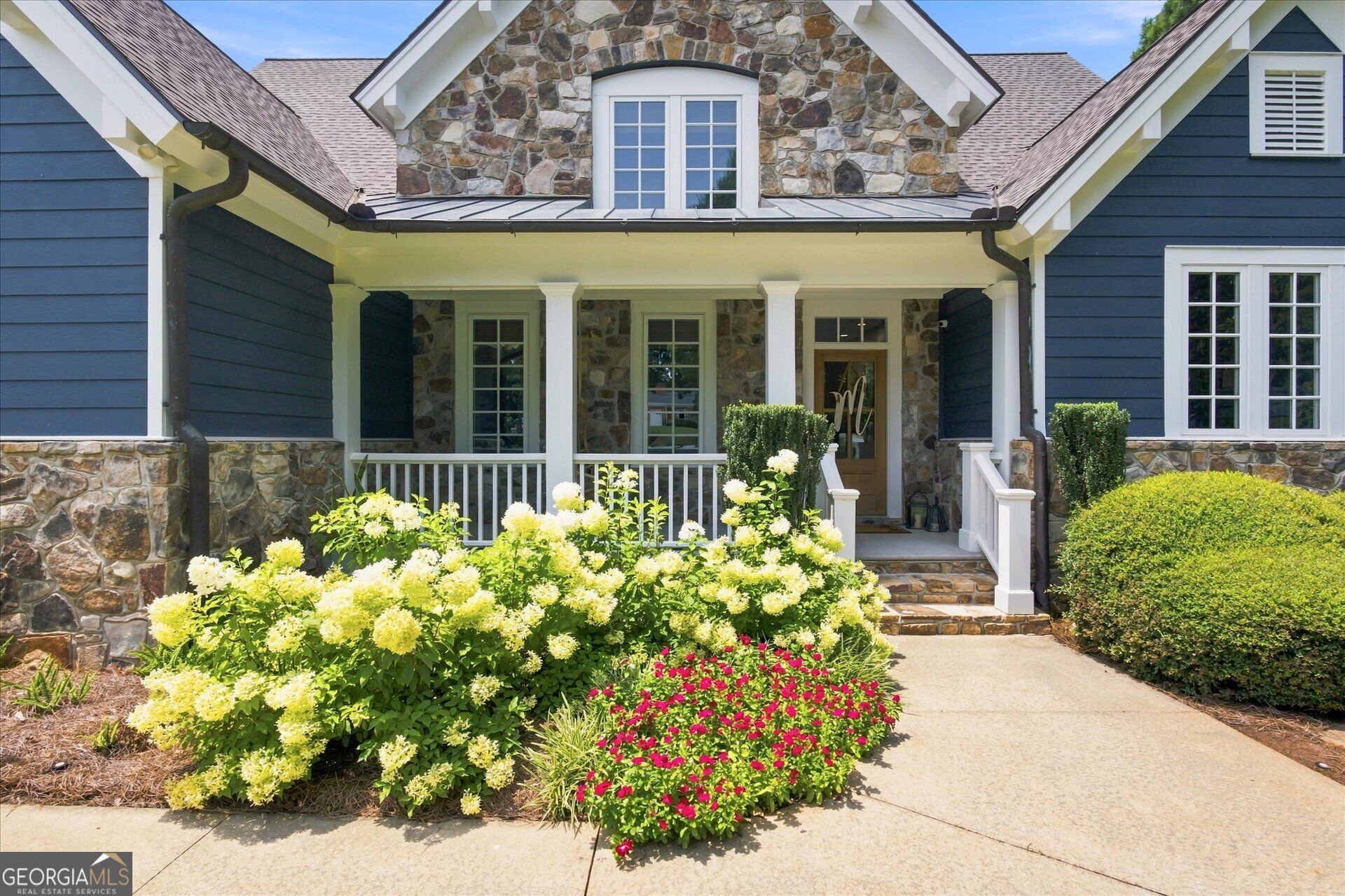 Home with a stone facade, blue siding, covered porch, and colorful flower beds.