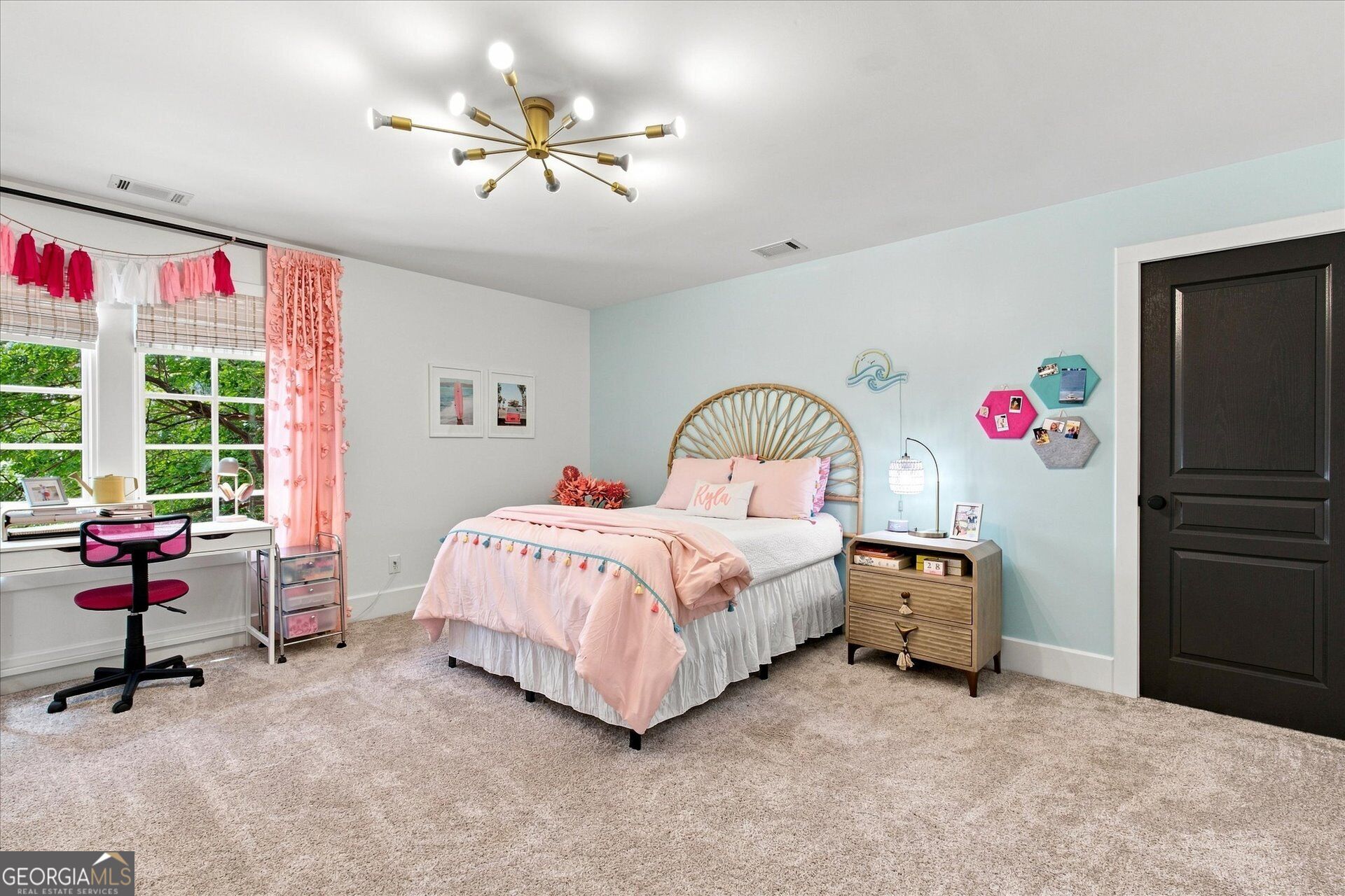 Bedroom with a pink bed, woven rattan headboard, pale blue wall, and desk beside large windows.