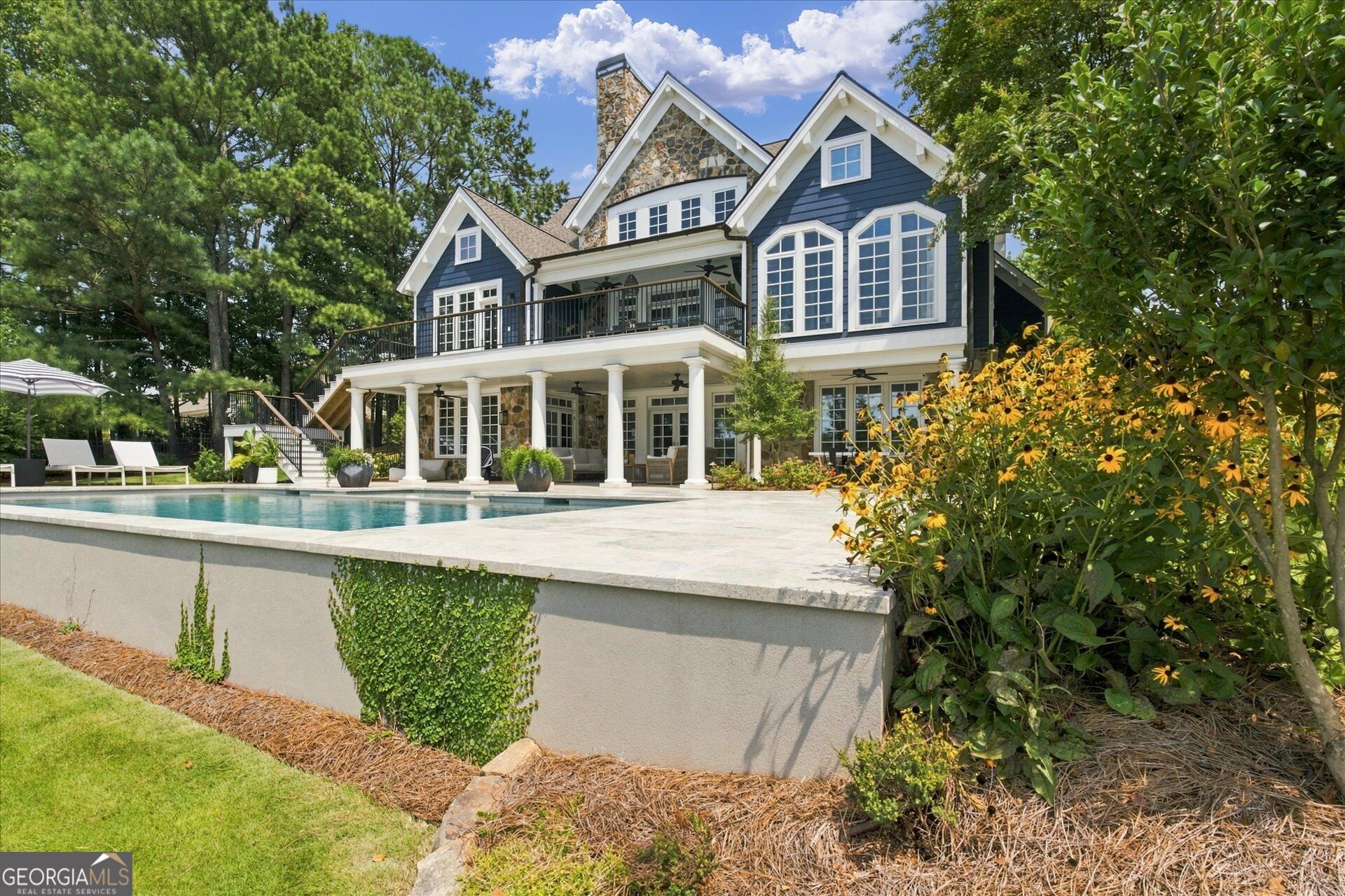 Blue-and-white home with stone accents, a covered patio, and swimming pool amid trees and flowering gardens.