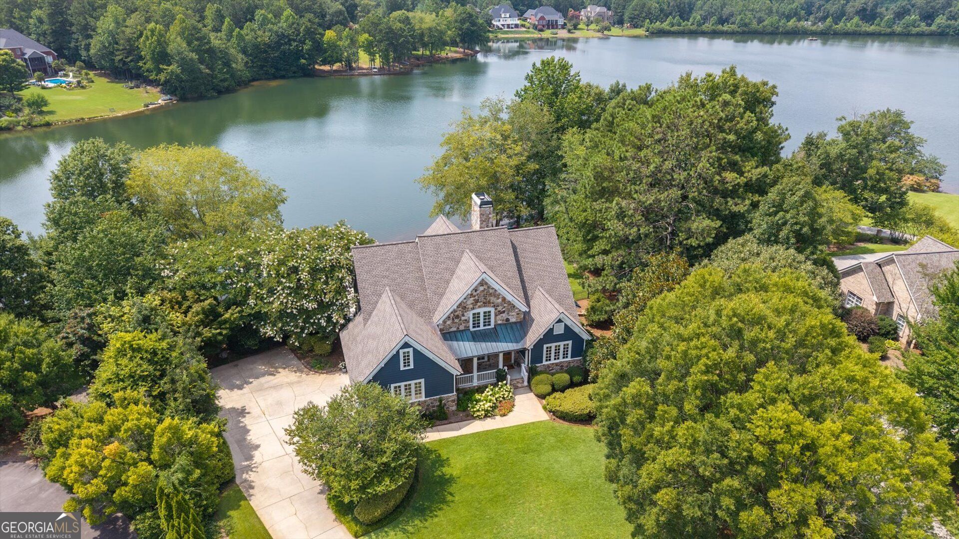 Aerial view of a blue and stone house beside a tree-lined lake, with a broad lawn and driveway.