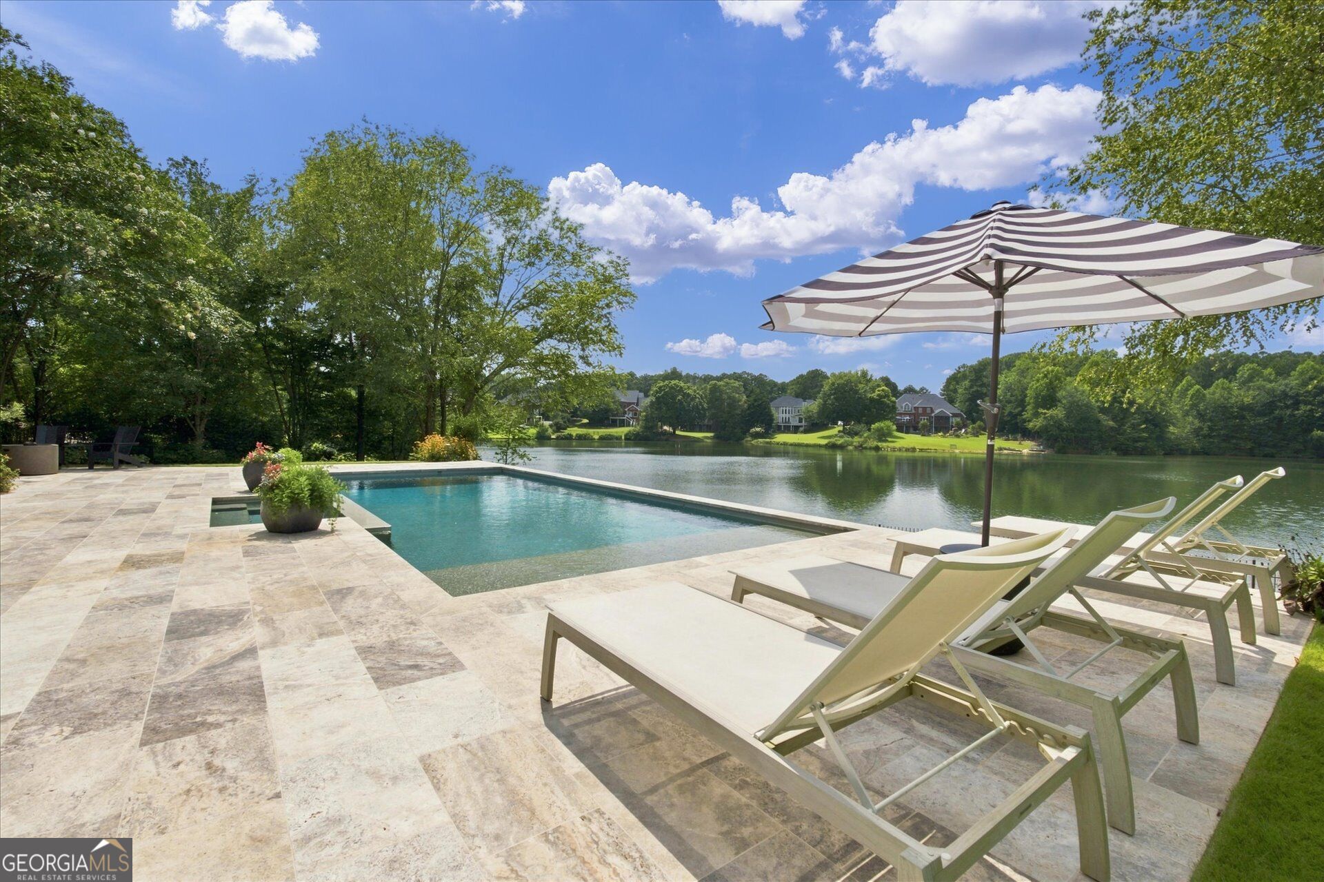 Lakeside pool and stone patio with lounge chairs beneath a striped umbrella.