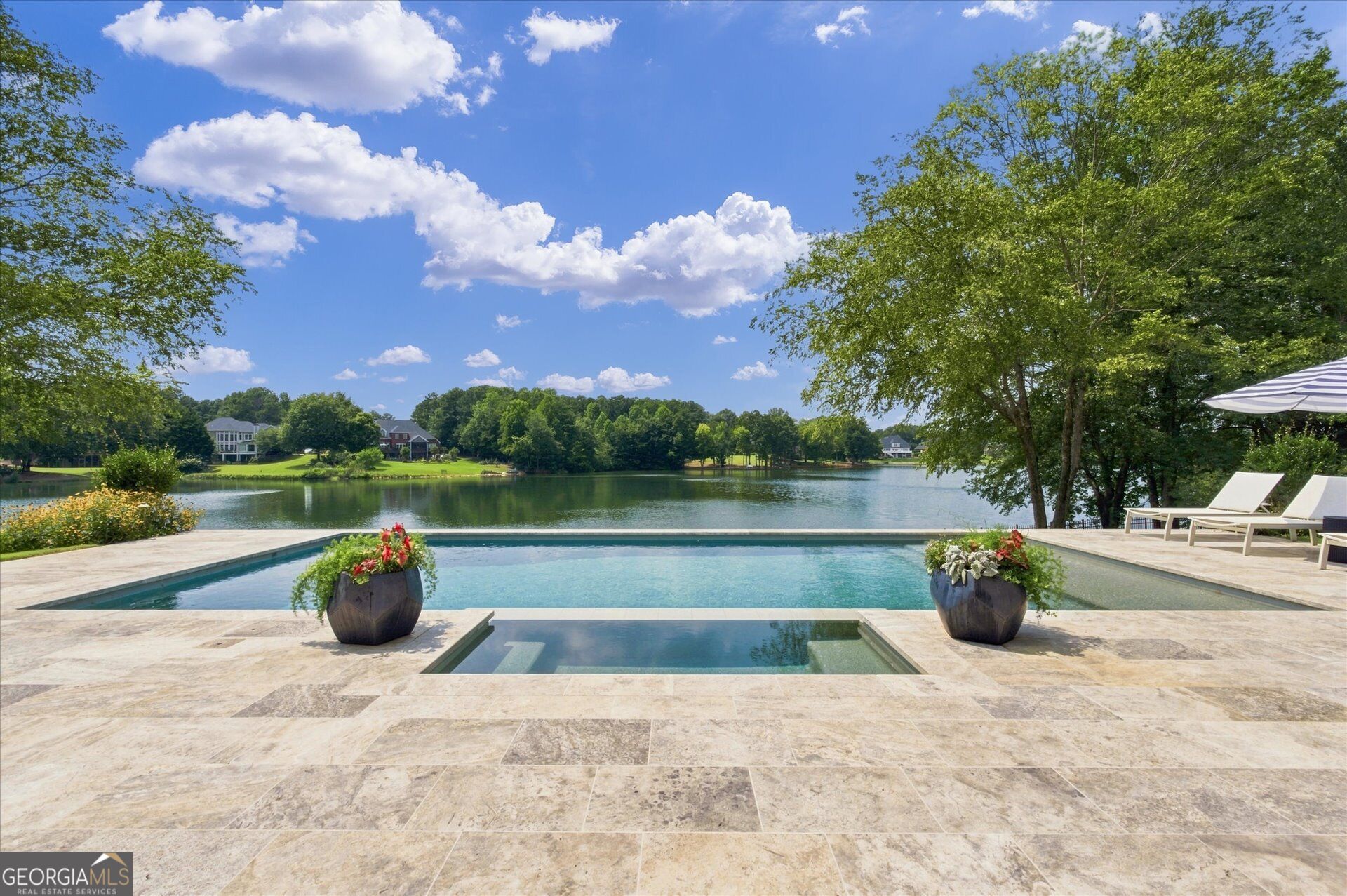 Swimming pool and spa overlook a tree-lined lake from a stone patio.