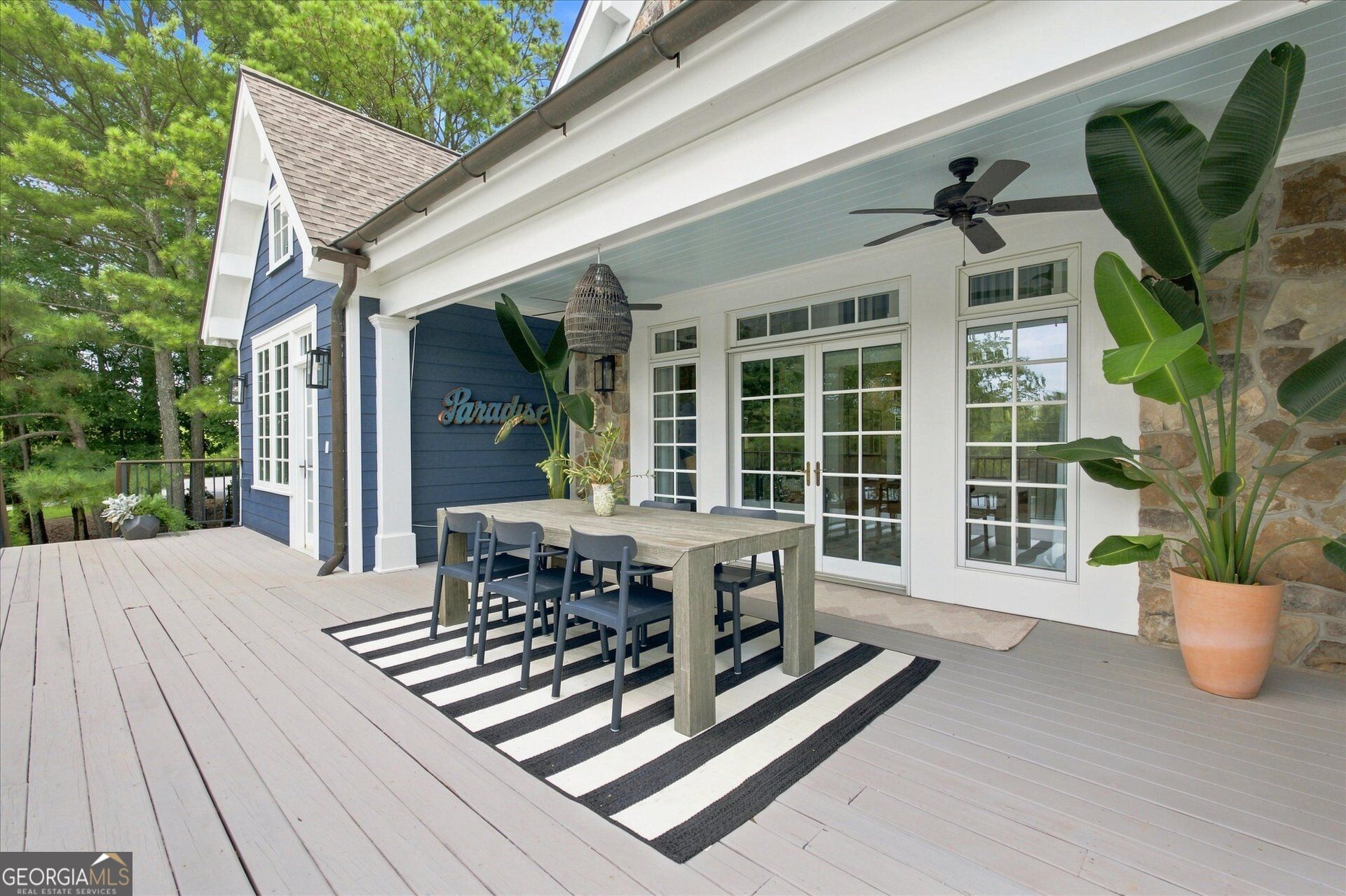 Covered deck with a dining table, striped rug, ceiling fan, and potted tropical plants.