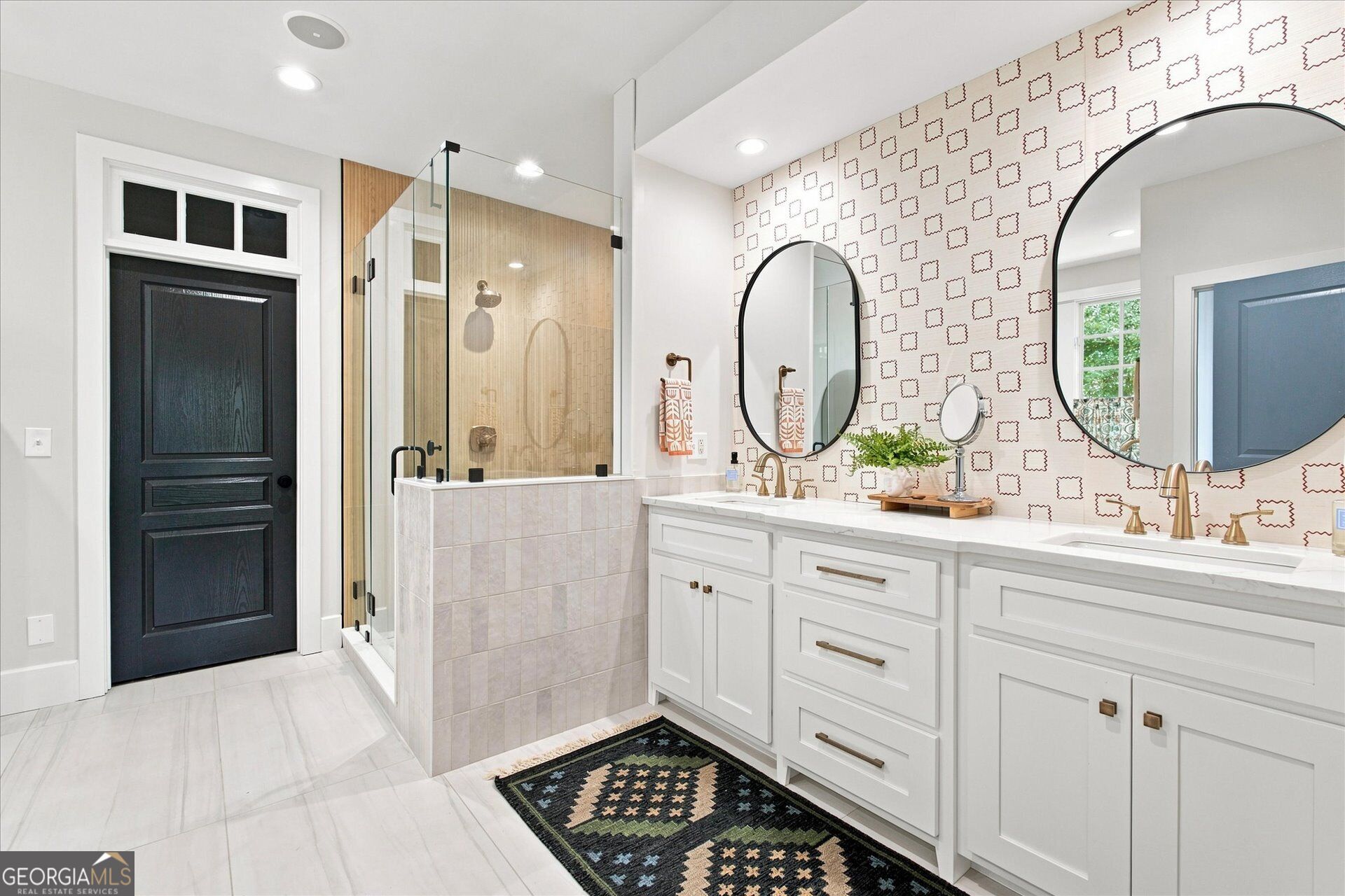 Bathroom with a glass shower, white double vanity, brass fixtures, and patterned wallpaper.