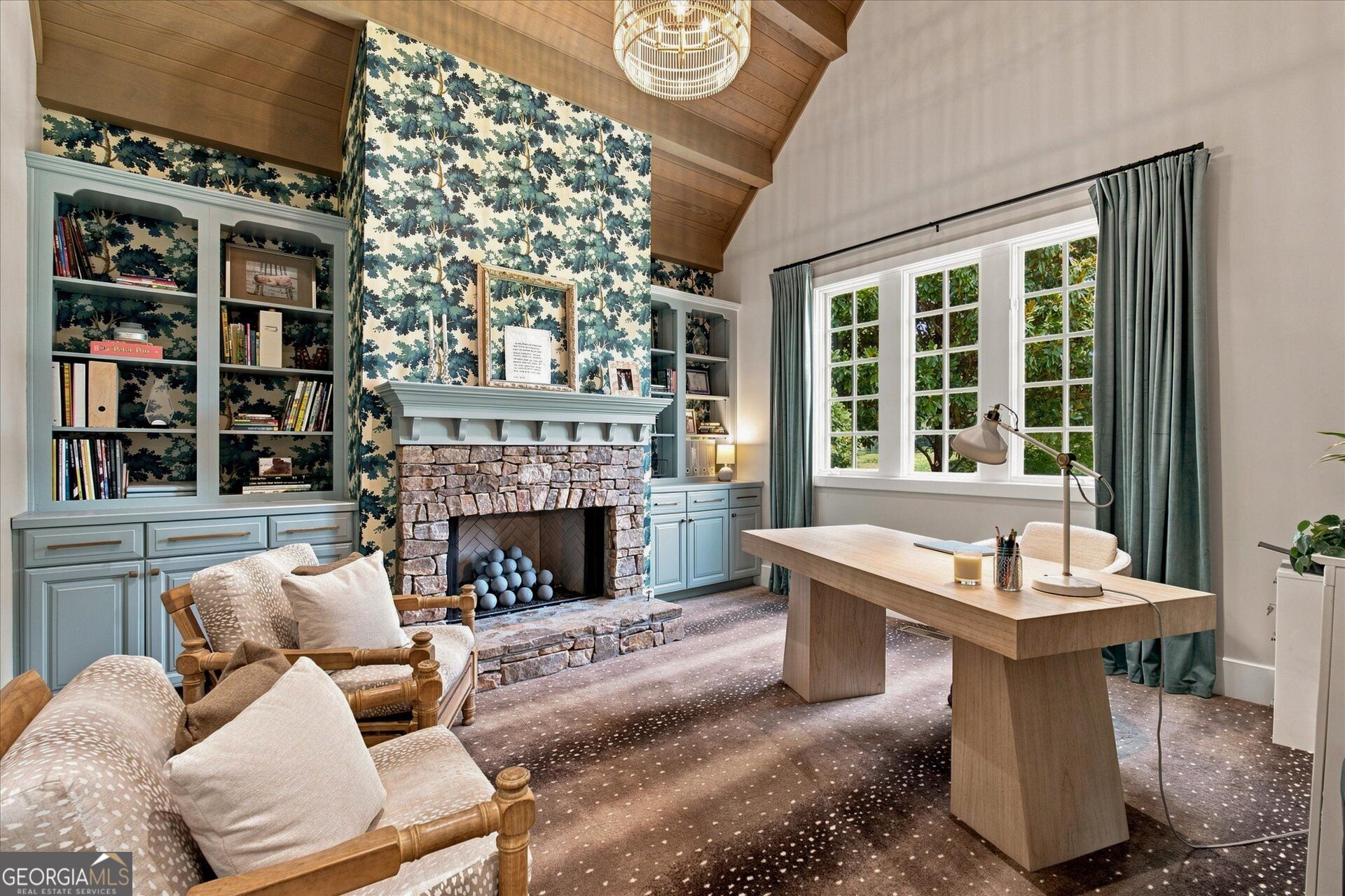 Study with vaulted wood ceiling, built-in bookshelves, stone fireplace, and desk by large windows.