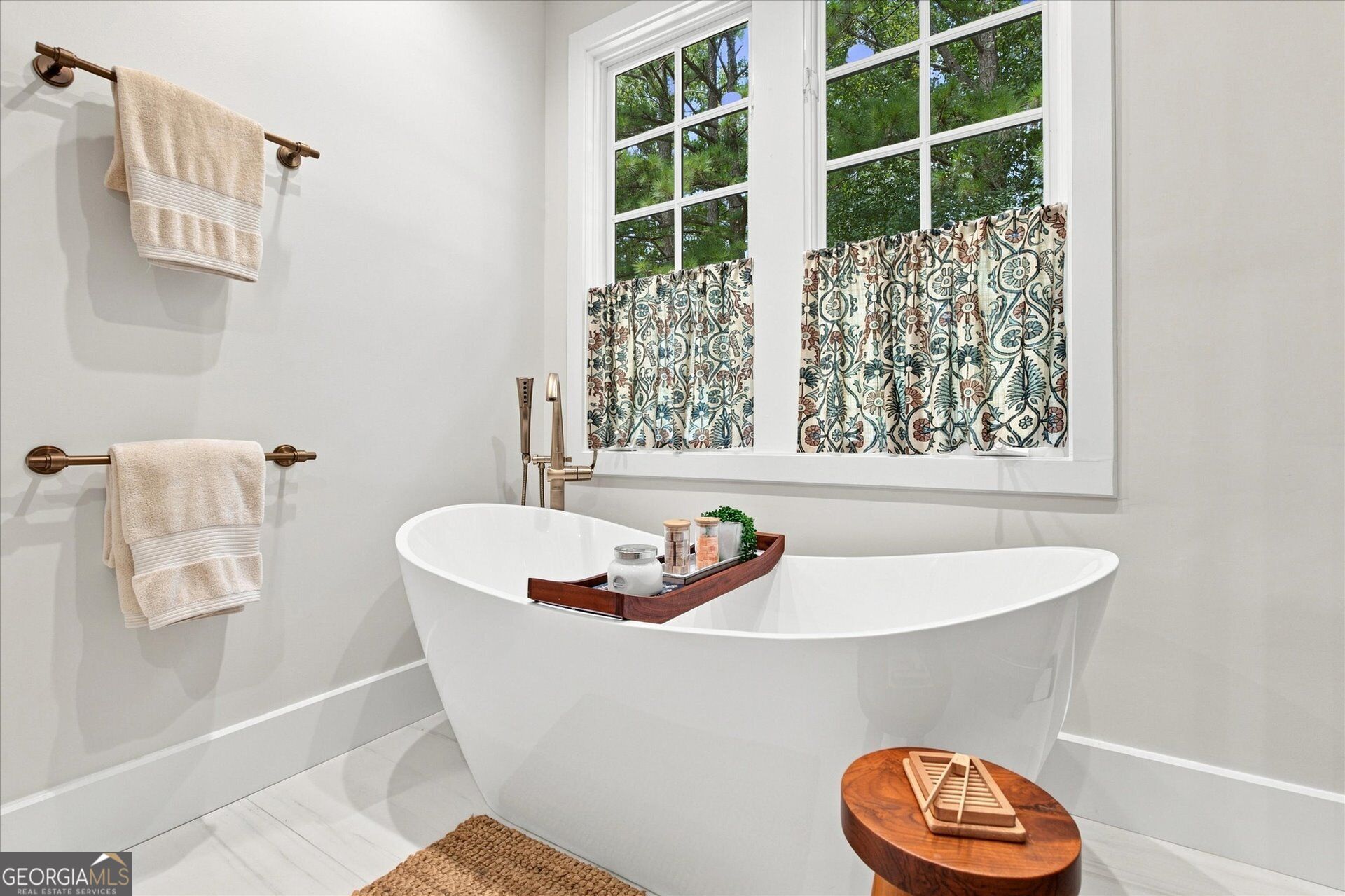 Bathroom with a freestanding soaking tub beneath large windows with patterned curtains.