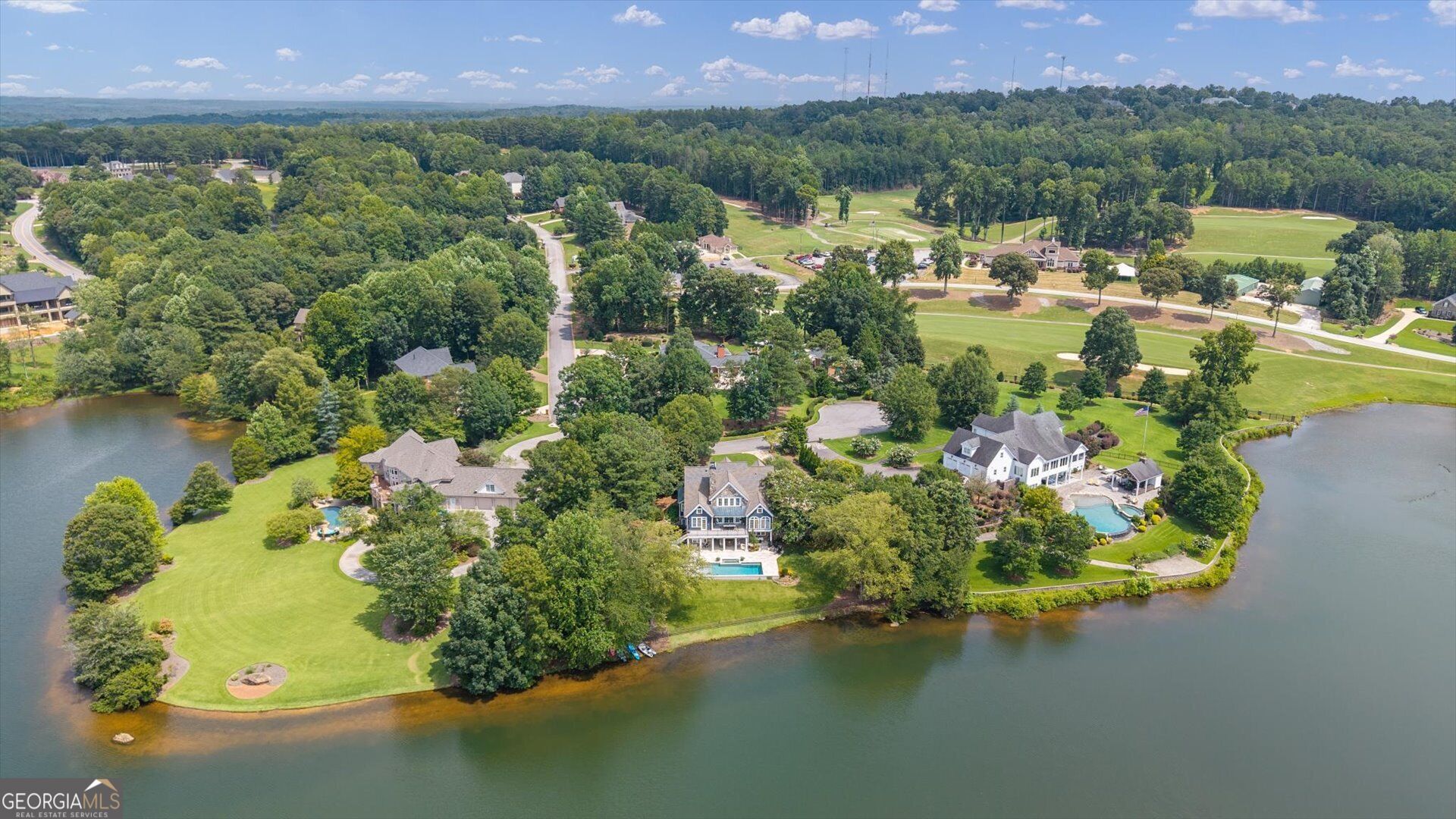 Aerial view of waterfront homes beside a lake, with pools, wooded grounds, and golf course greens.
