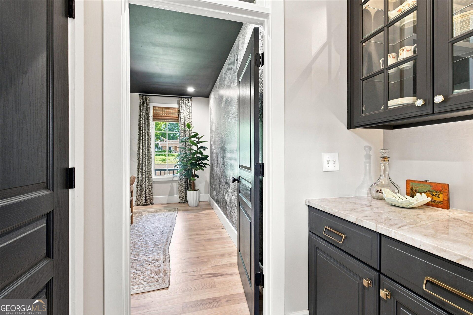 Hallway with light wood floors, a dark ceiling, patterned accent wall, and a potted plant by the window.
