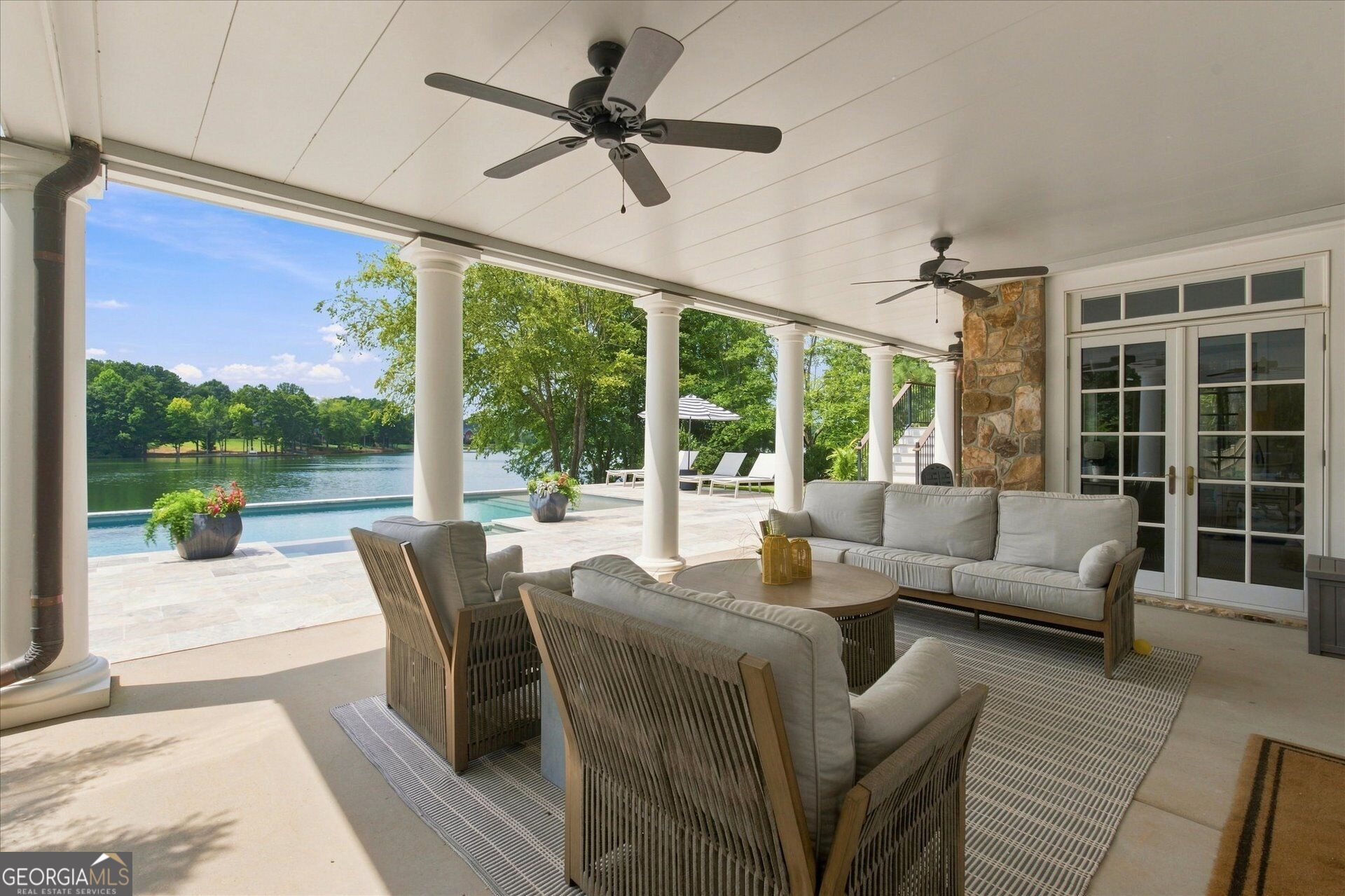 Covered patio with cushioned seating overlooks a swimming pool and tree-lined lake.