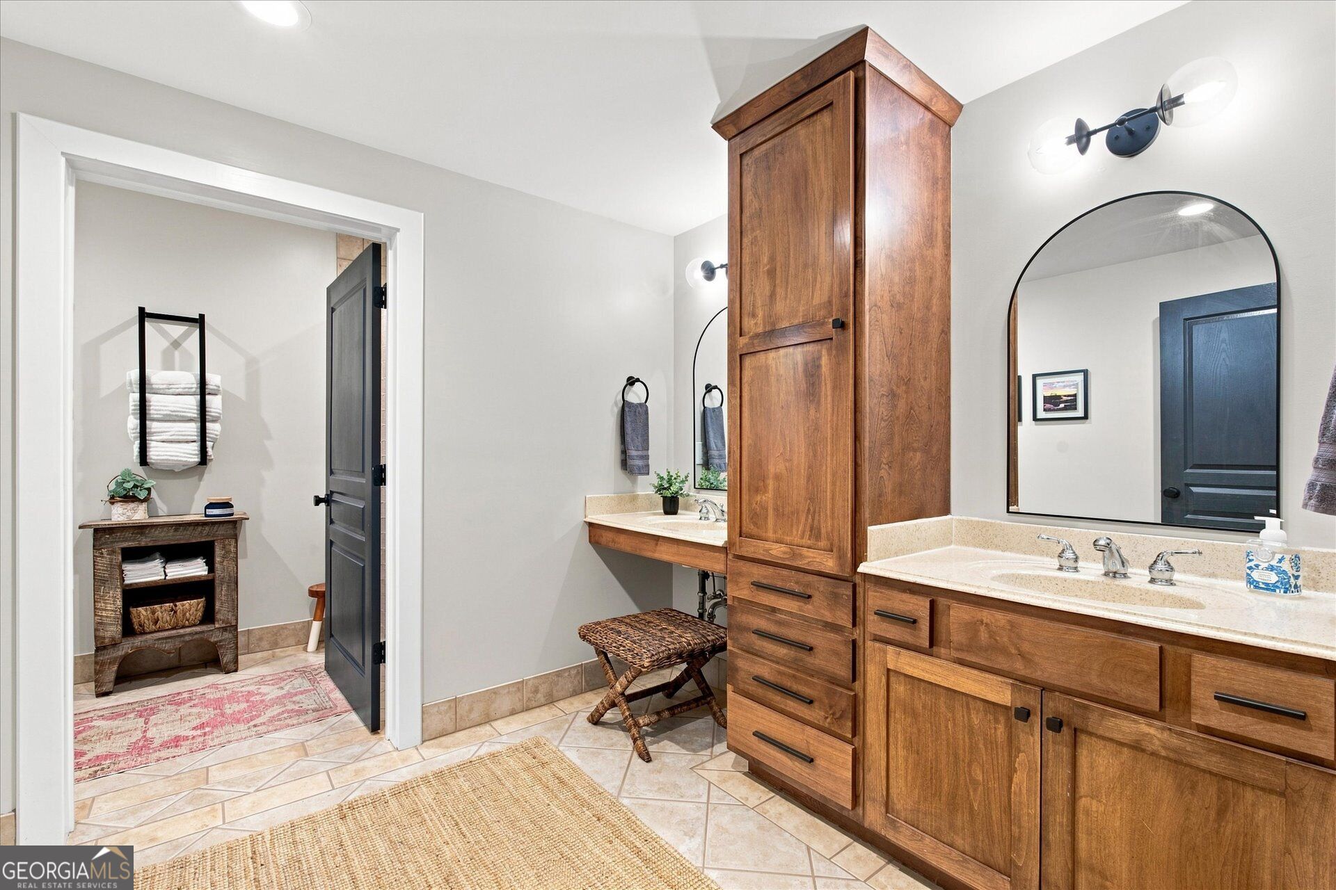 Bathroom with a long wood vanity, tall central cabinet, arched mirror, and tiled floor.