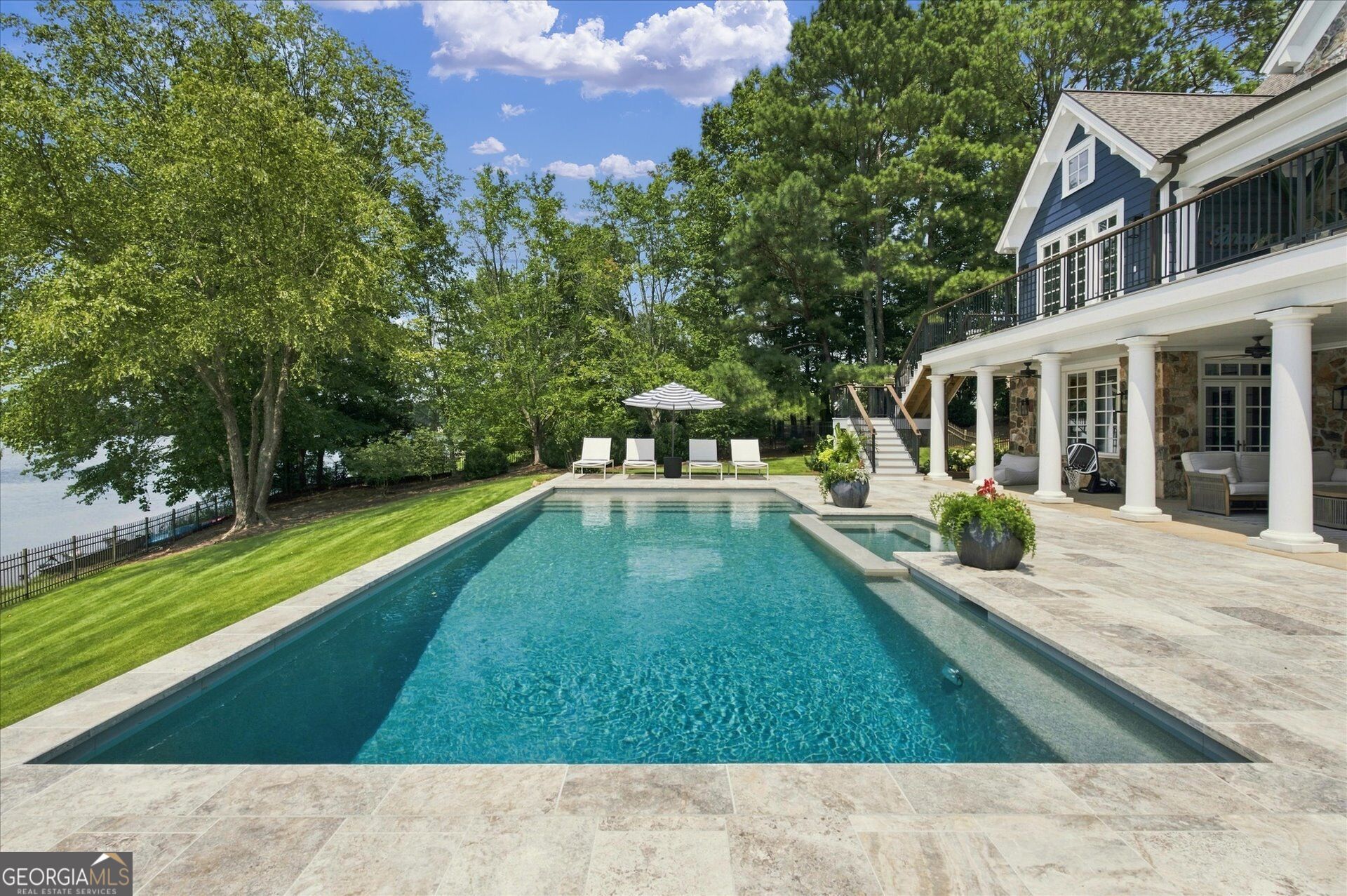 Backyard pool beside a blue-and-white home, with a covered terrace, stone patio, and wooded water view.