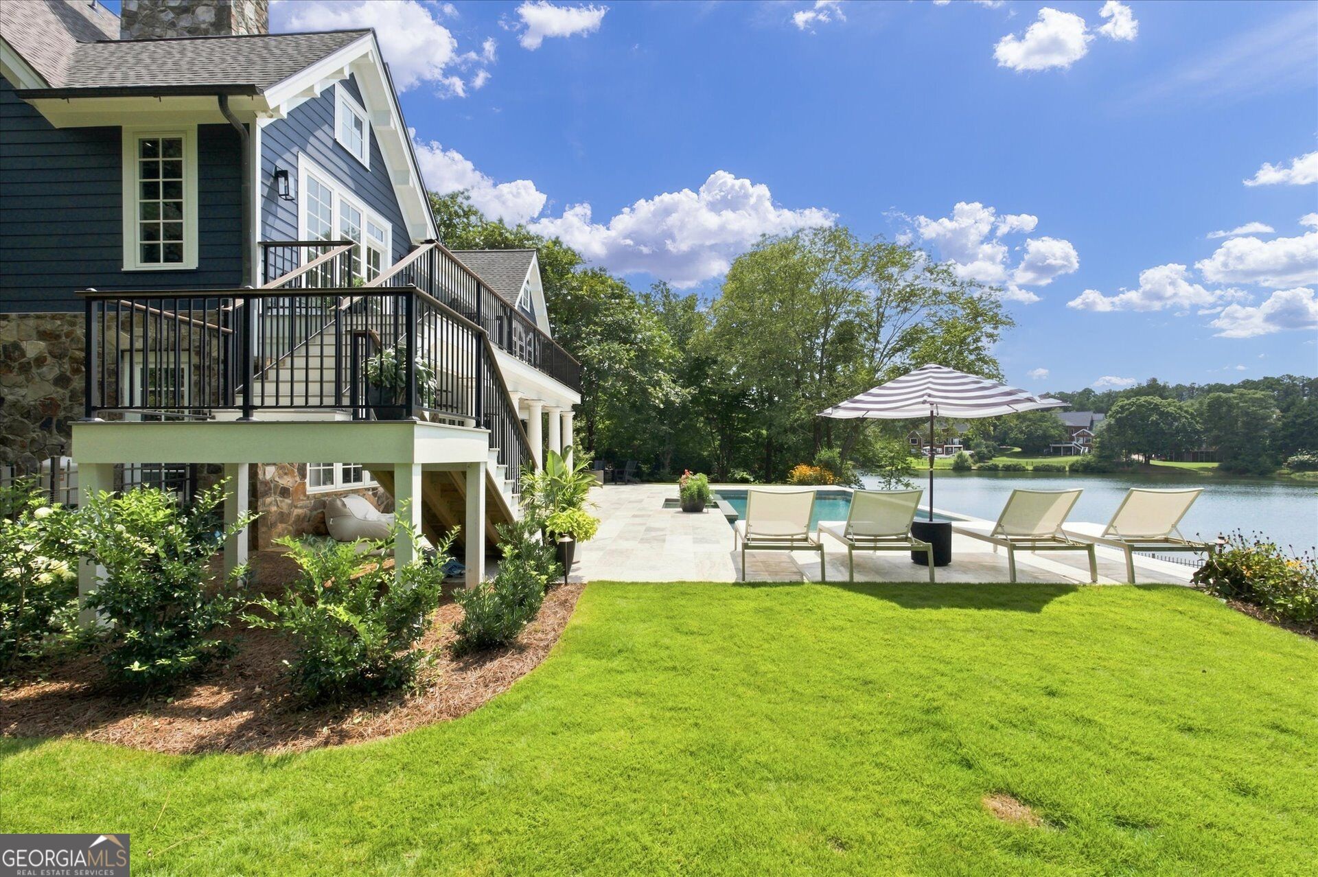 Dark blue house with a poolside patio, lounge chairs, and a striped umbrella overlooking a lake.