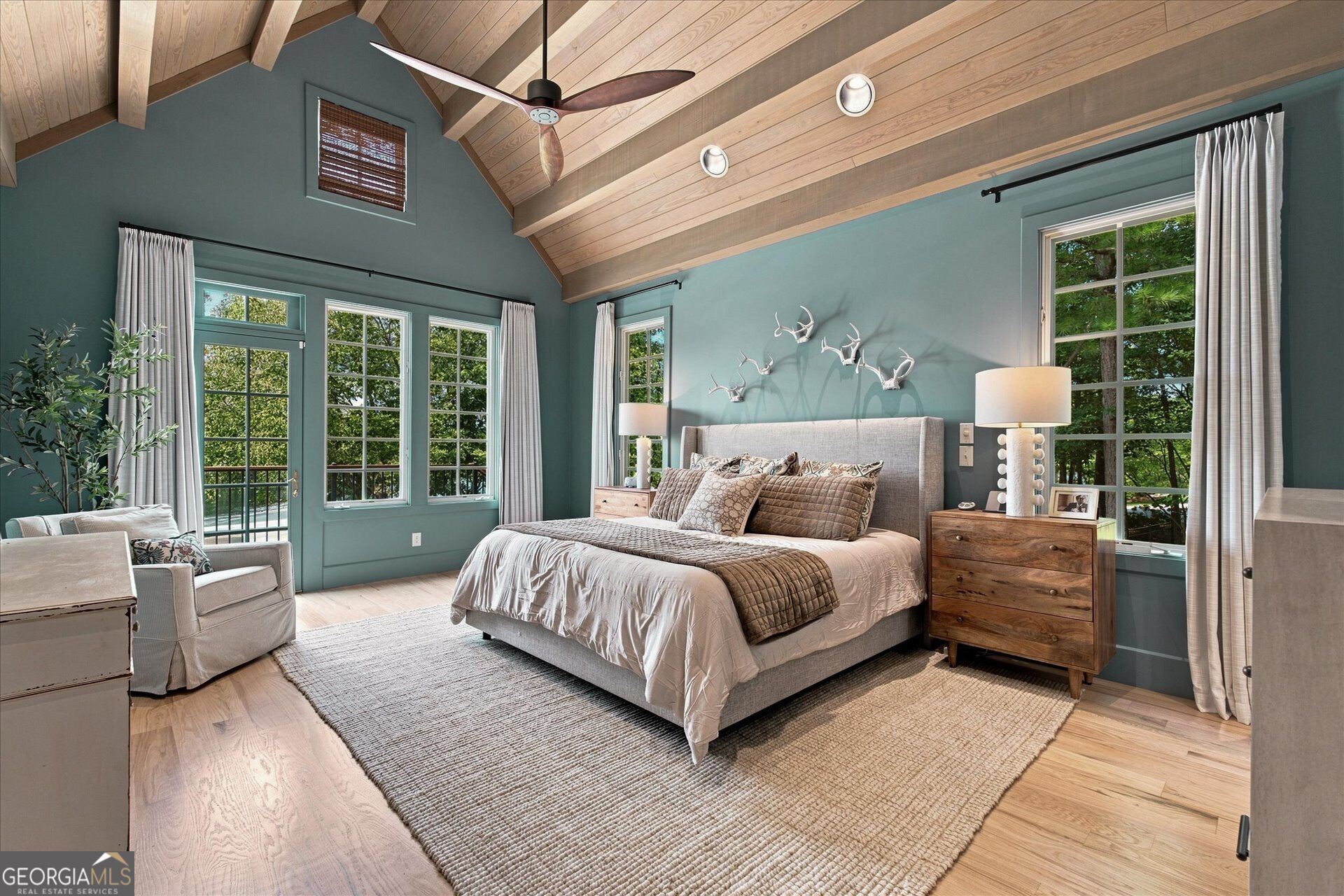 Furnished bedroom with a vaulted wood ceiling, teal walls, and tall windows overlooking trees.