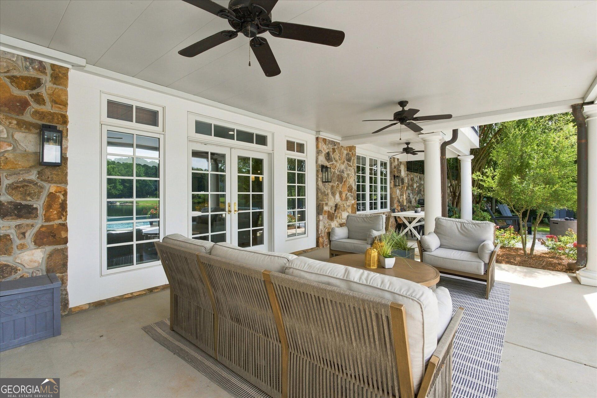 Covered patio with stone walls, ceiling fans, and cushioned seating beside a lake.
