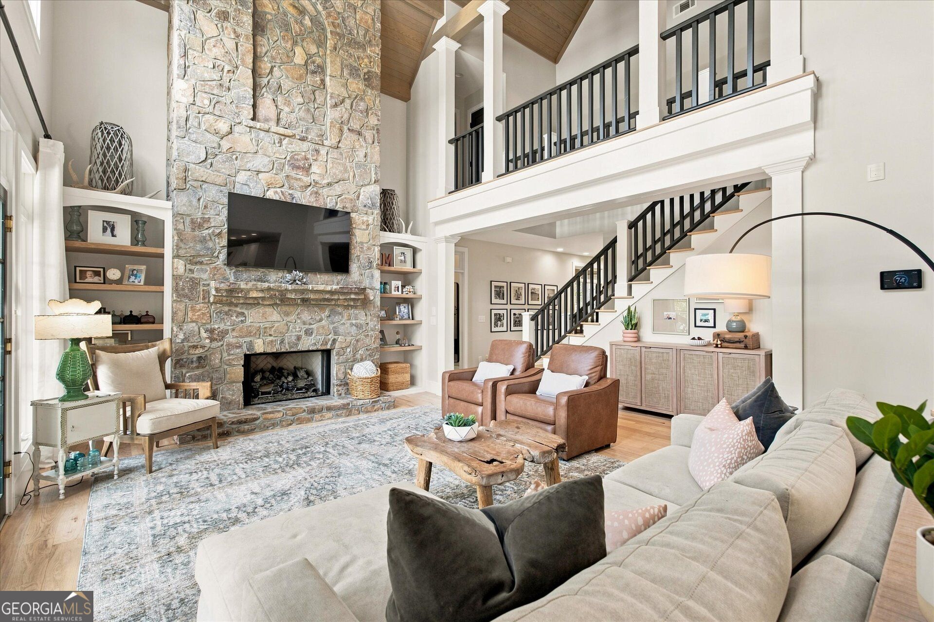 Living room with a tall stone fireplace, vaulted wood ceiling, and open staircase.