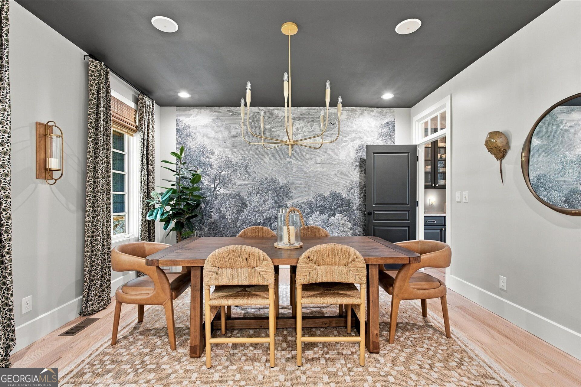 Dining room with a wood table, woven chairs, brass chandelier, and monochrome landscape mural.