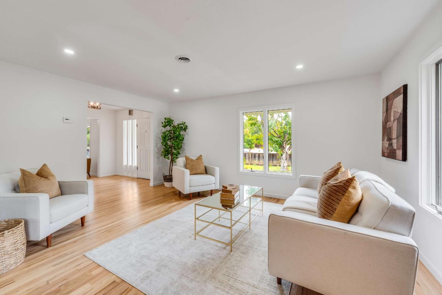 Staged living room with white seating, tan pillows, a glass coffee table, and windows overlooking a leafy yard.