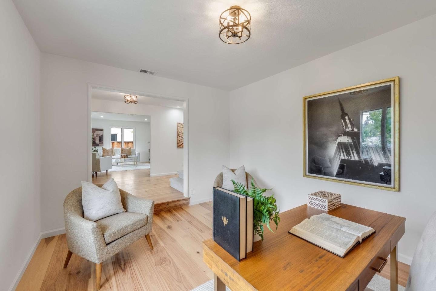 Staged home office with a wooden desk, open book, plant, and doorway to the living room.