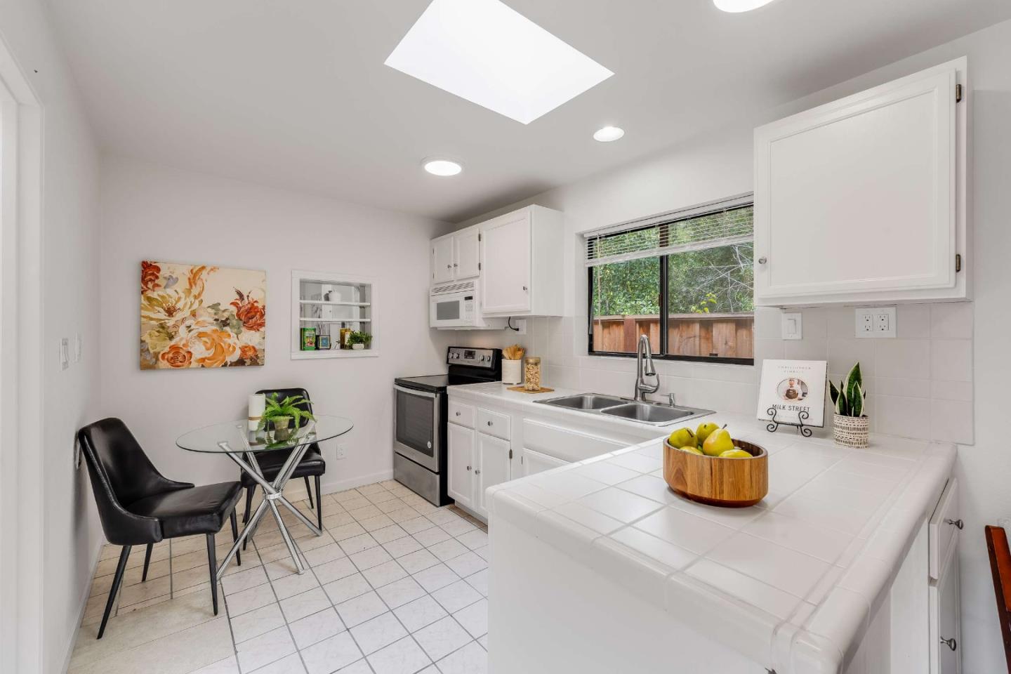 White kitchen with tiled counters, a double sink beneath a window, and a small glass-table dining nook.