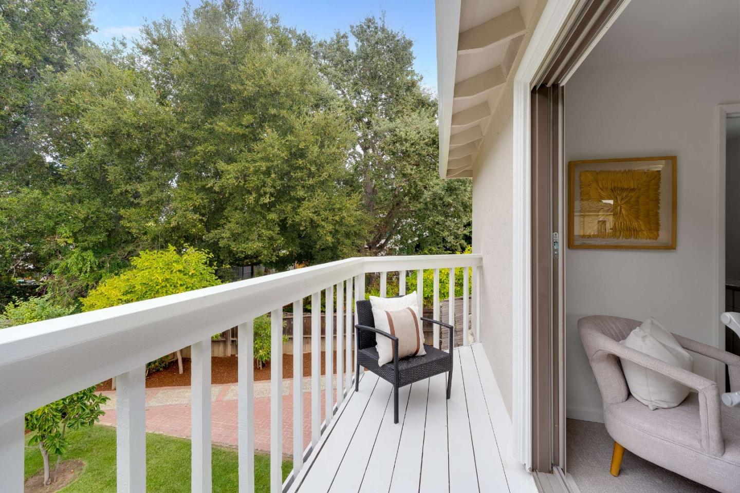 White balcony overlooking a leafy yard, with a black chair beside an open sliding glass door.