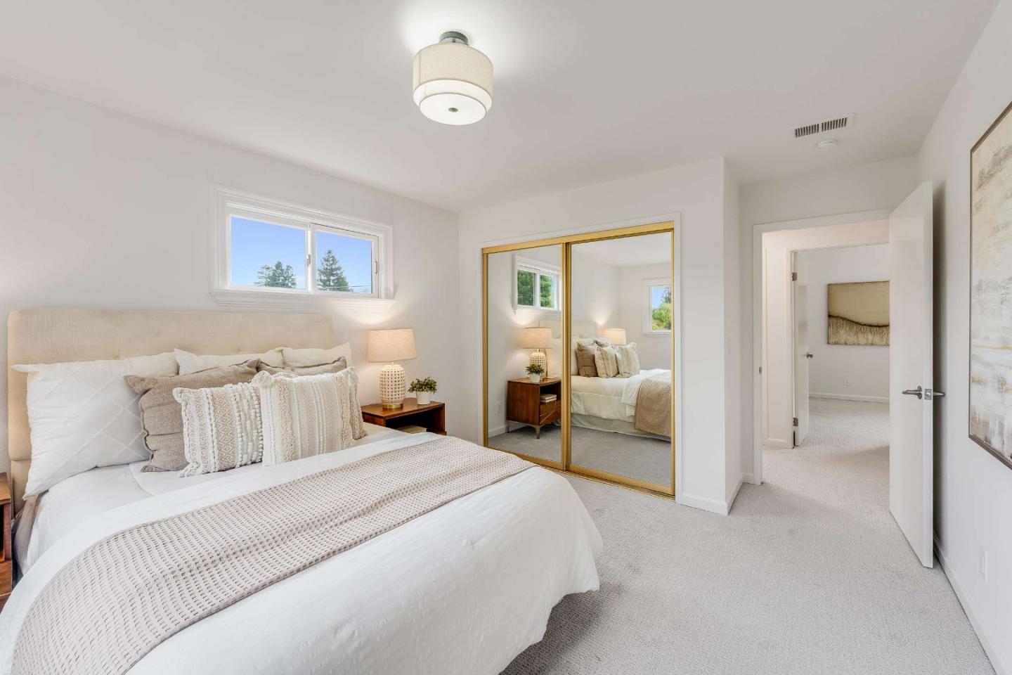 Bright bedroom with a white bed, gold-framed mirrored closet doors, and a window above the headboard.