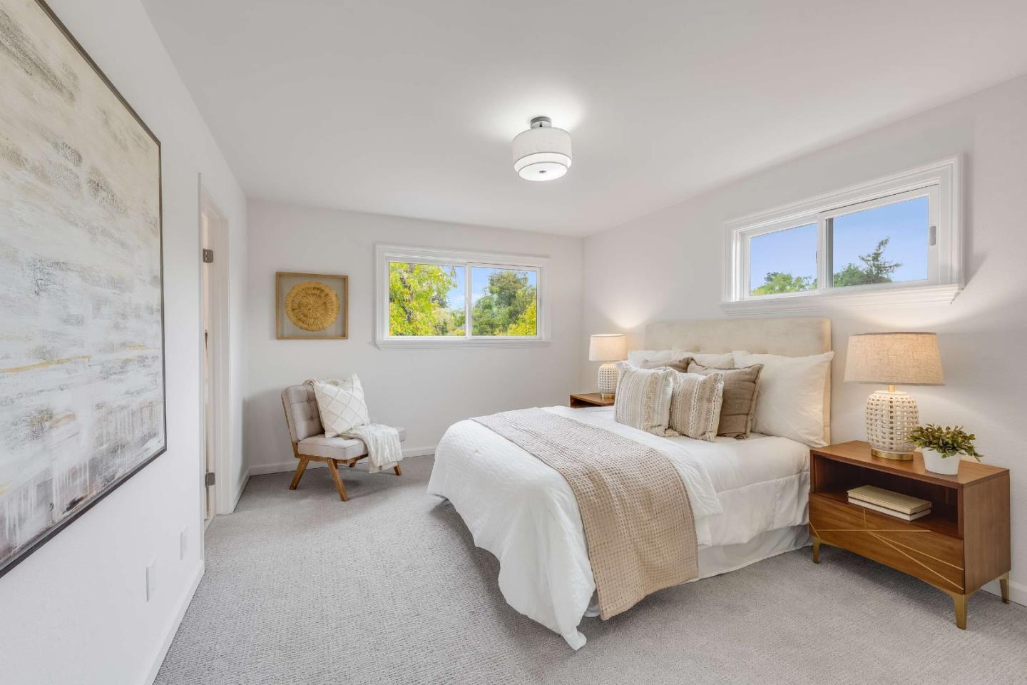 Staged bedroom with a white bed, wood nightstands, a small armchair, and windows overlooking trees.