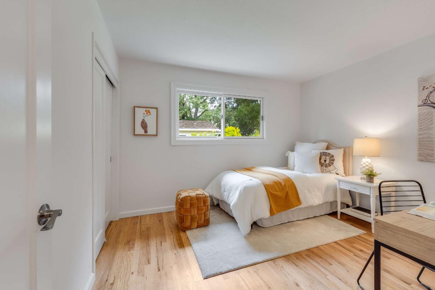 Staged bedroom with a white bed, mustard throw, woven ottoman, and window overlooking greenery.
