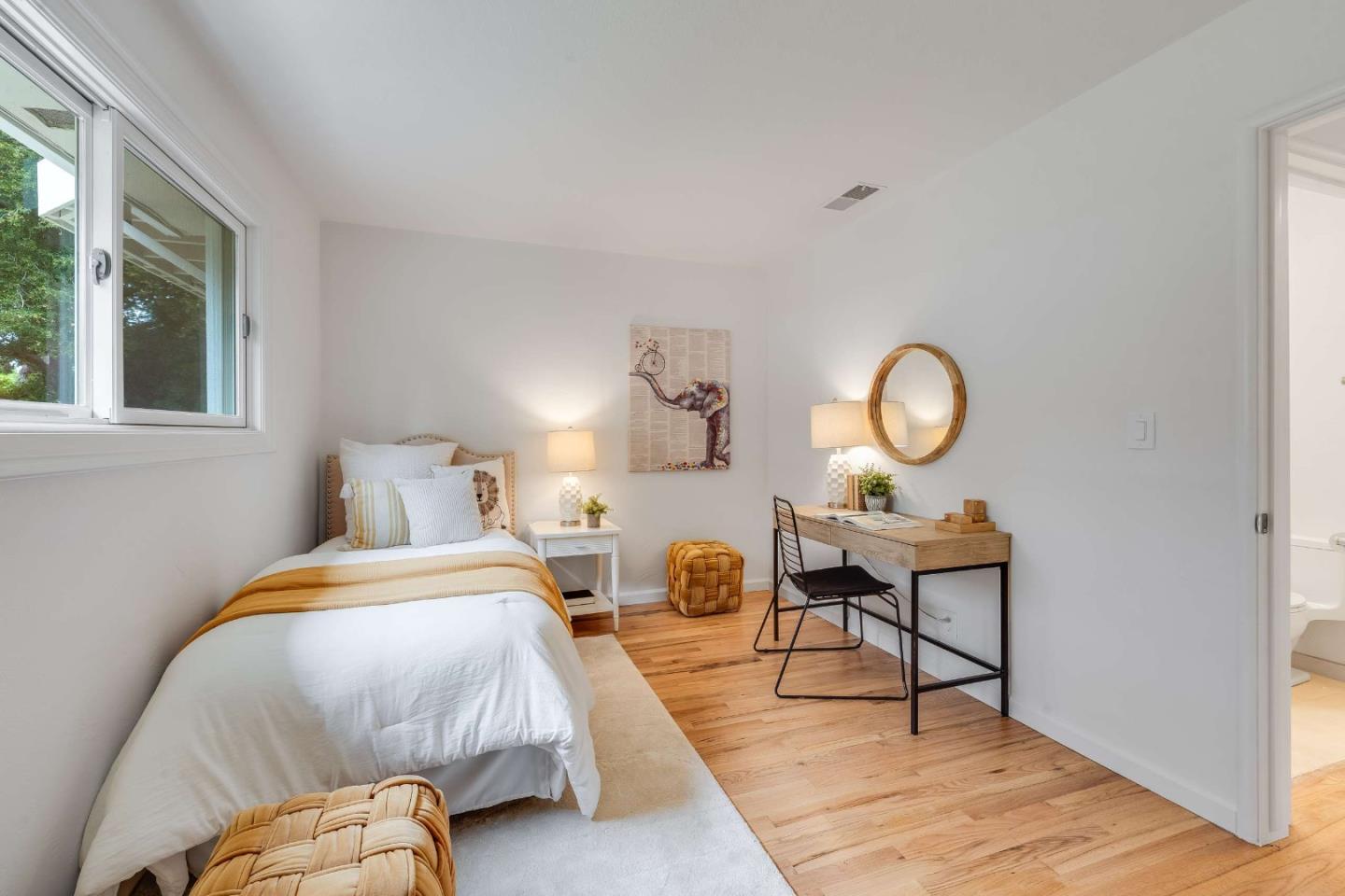 Staged bedroom with a single bed, wooden desk, round mirror, and hardwood floors.
