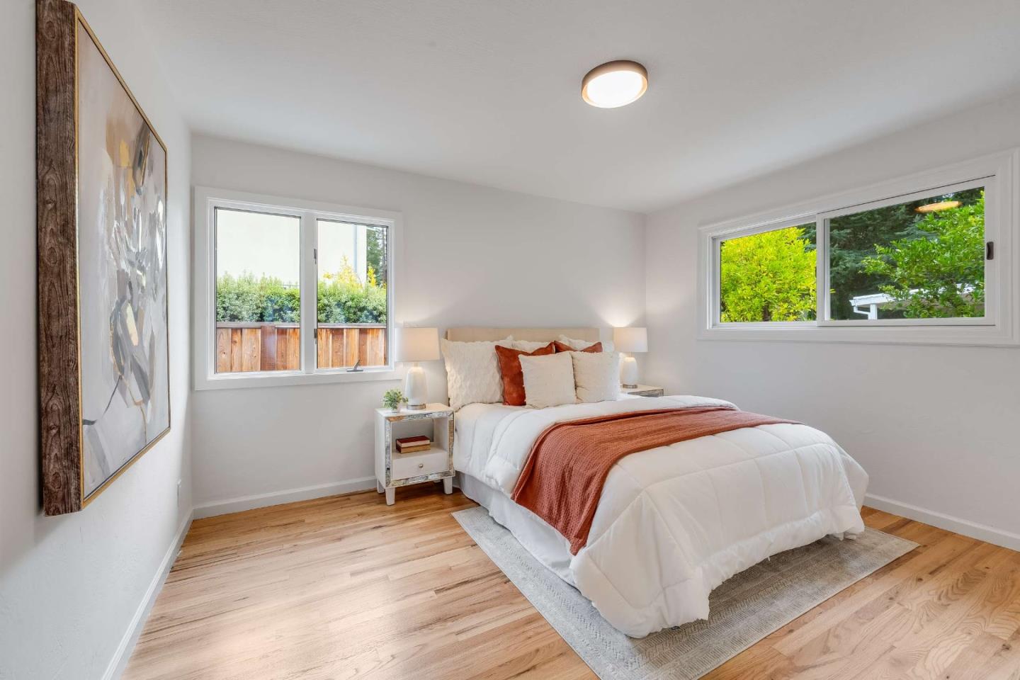 Staged bedroom with a white bed, rust-colored accents, wood floors, and windows overlooking greenery.