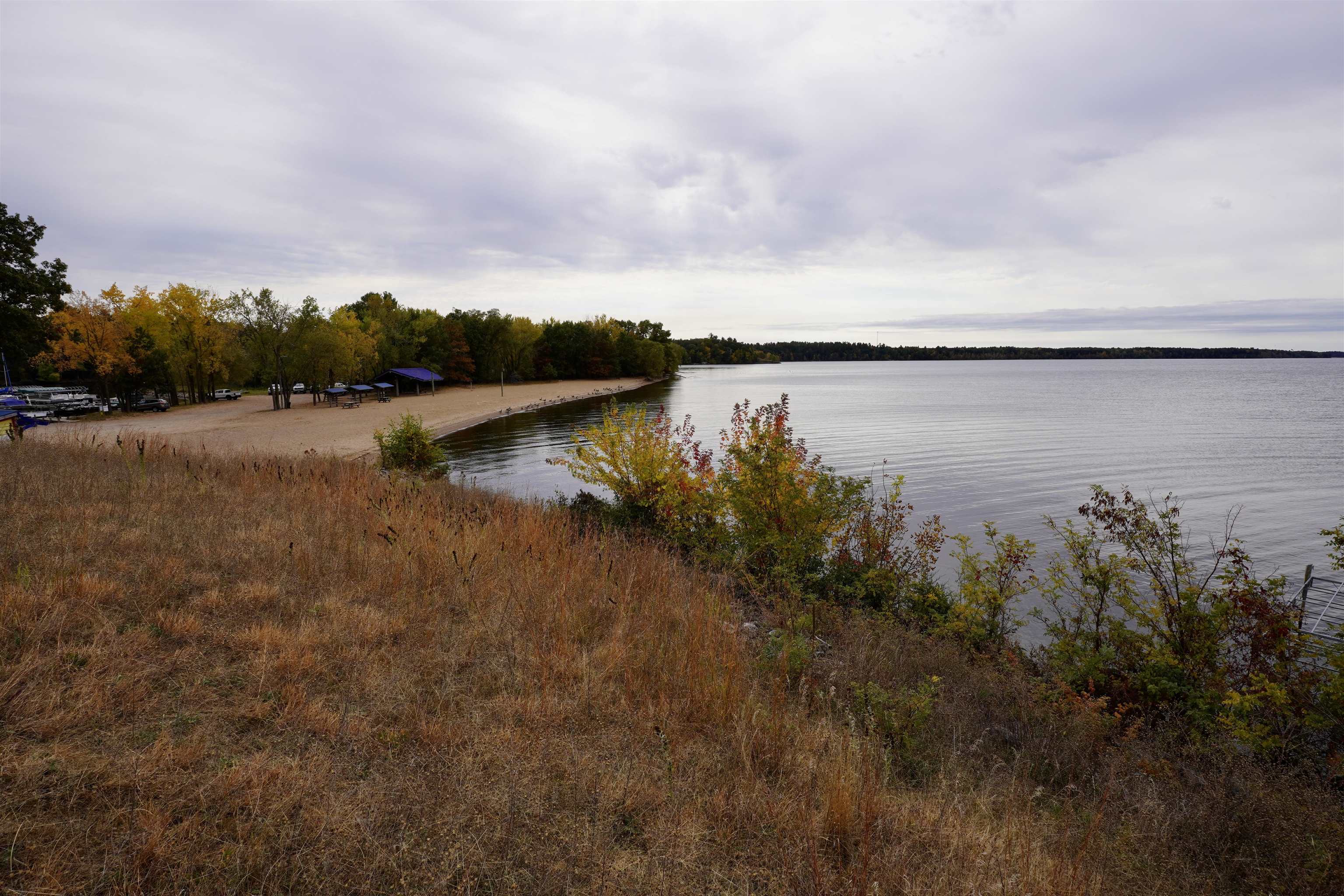 Sandy lakeshore beach with autumn trees, picnic tables, and a covered shelter beside calm water.