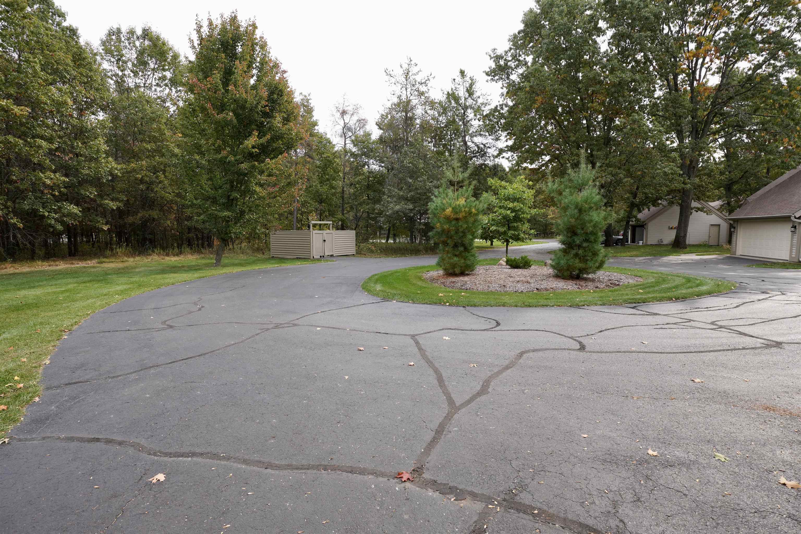 Curving paved driveway around a landscaped island, with wooded grounds and beige garages nearby.
