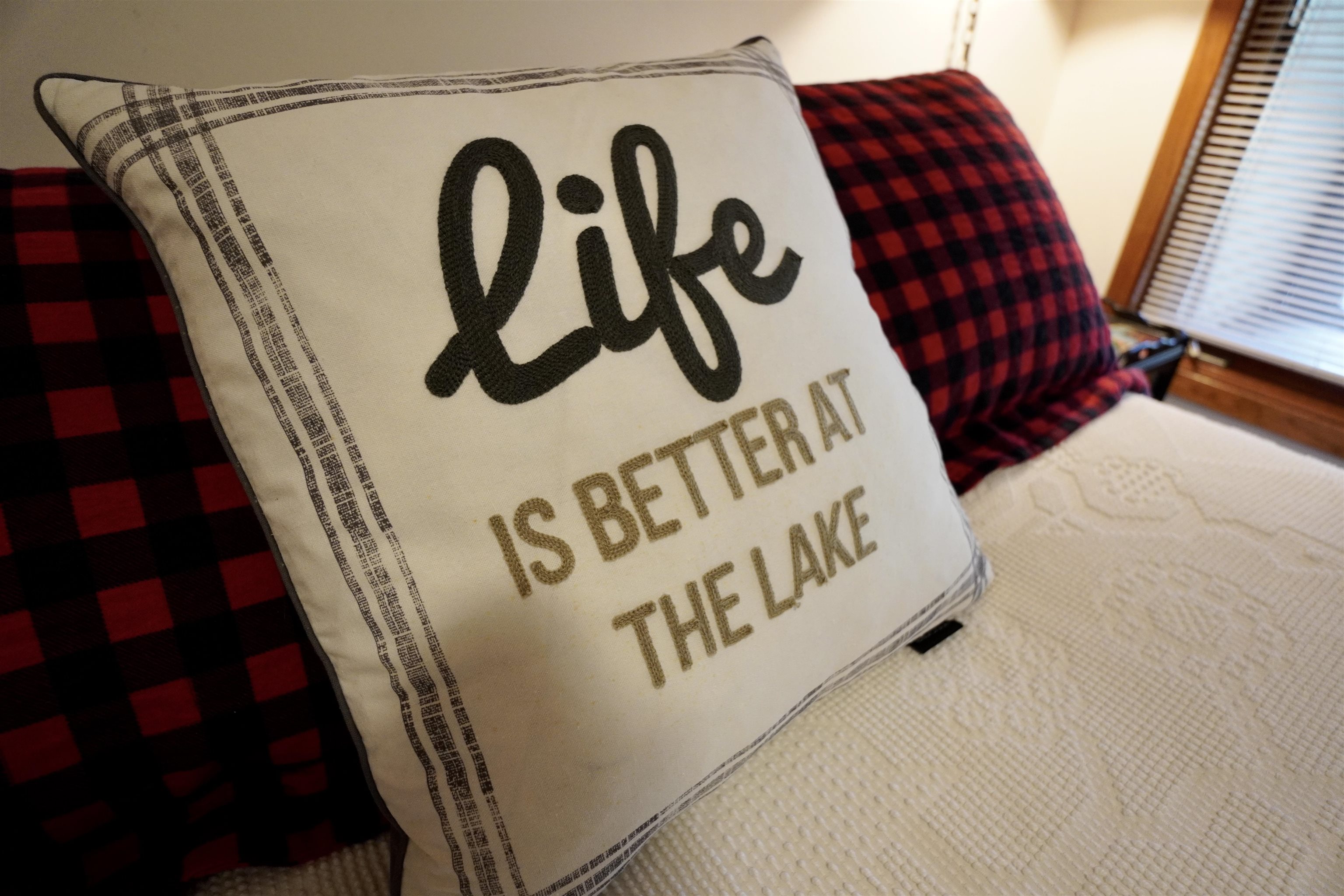Lake-themed embroidered pillow on a white bed beside red-and-black check pillows.