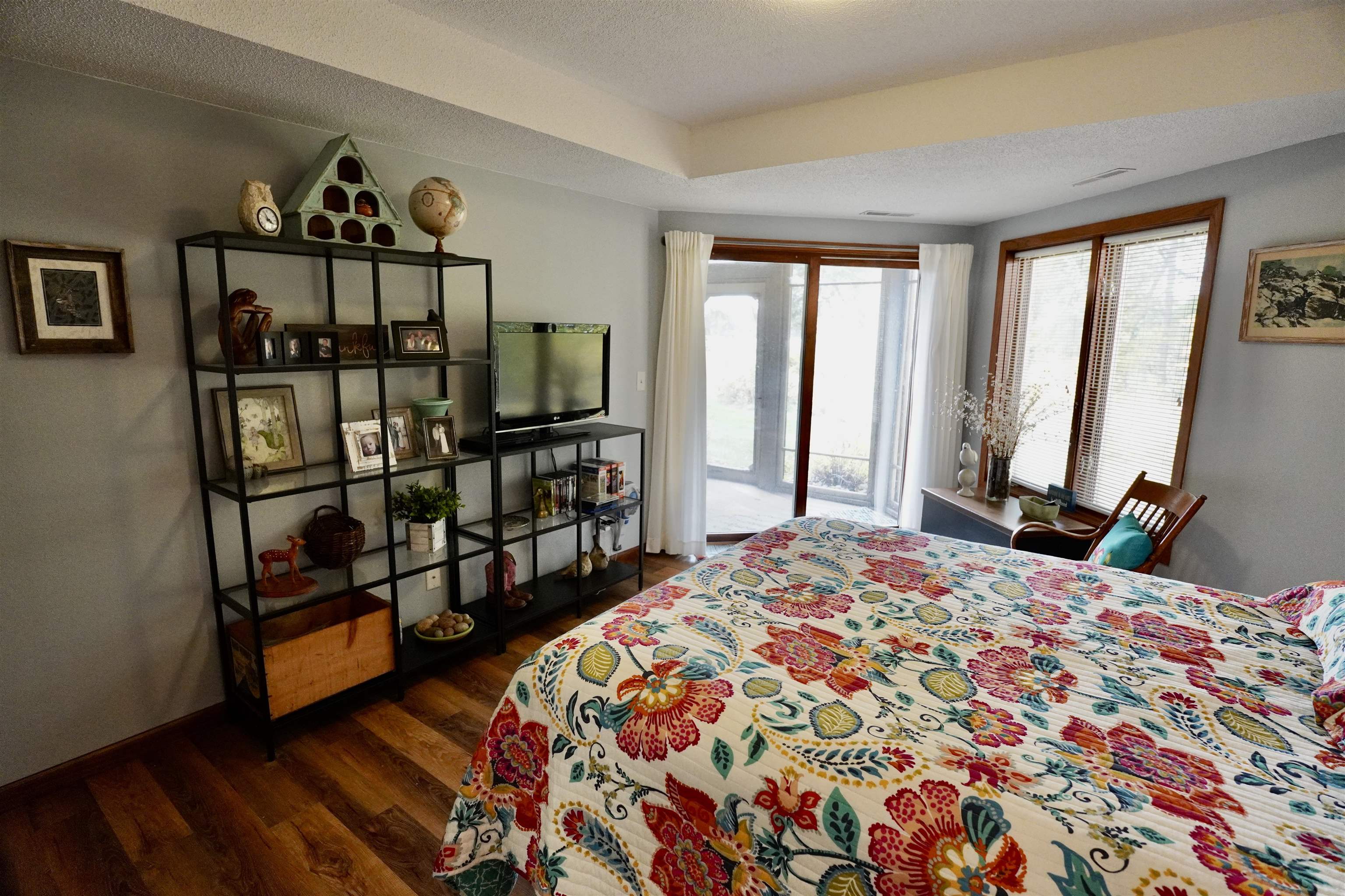 Bedroom with a colorful floral quilt, large windows, and open shelving beside the bed.