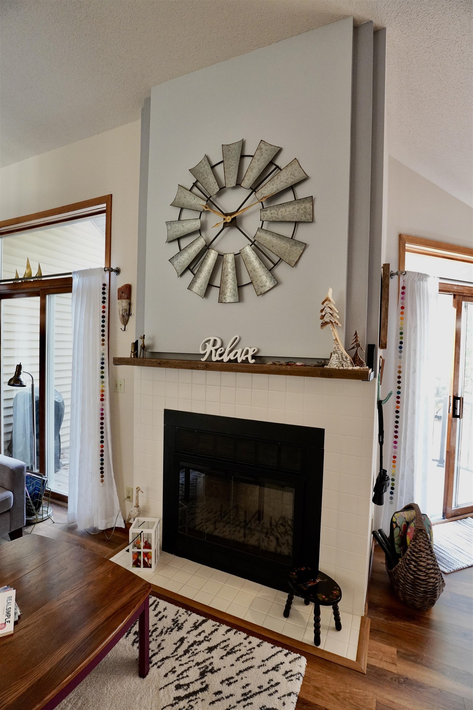 Living room fireplace with a white tiled surround, oversized metal clock, and windows on either side.