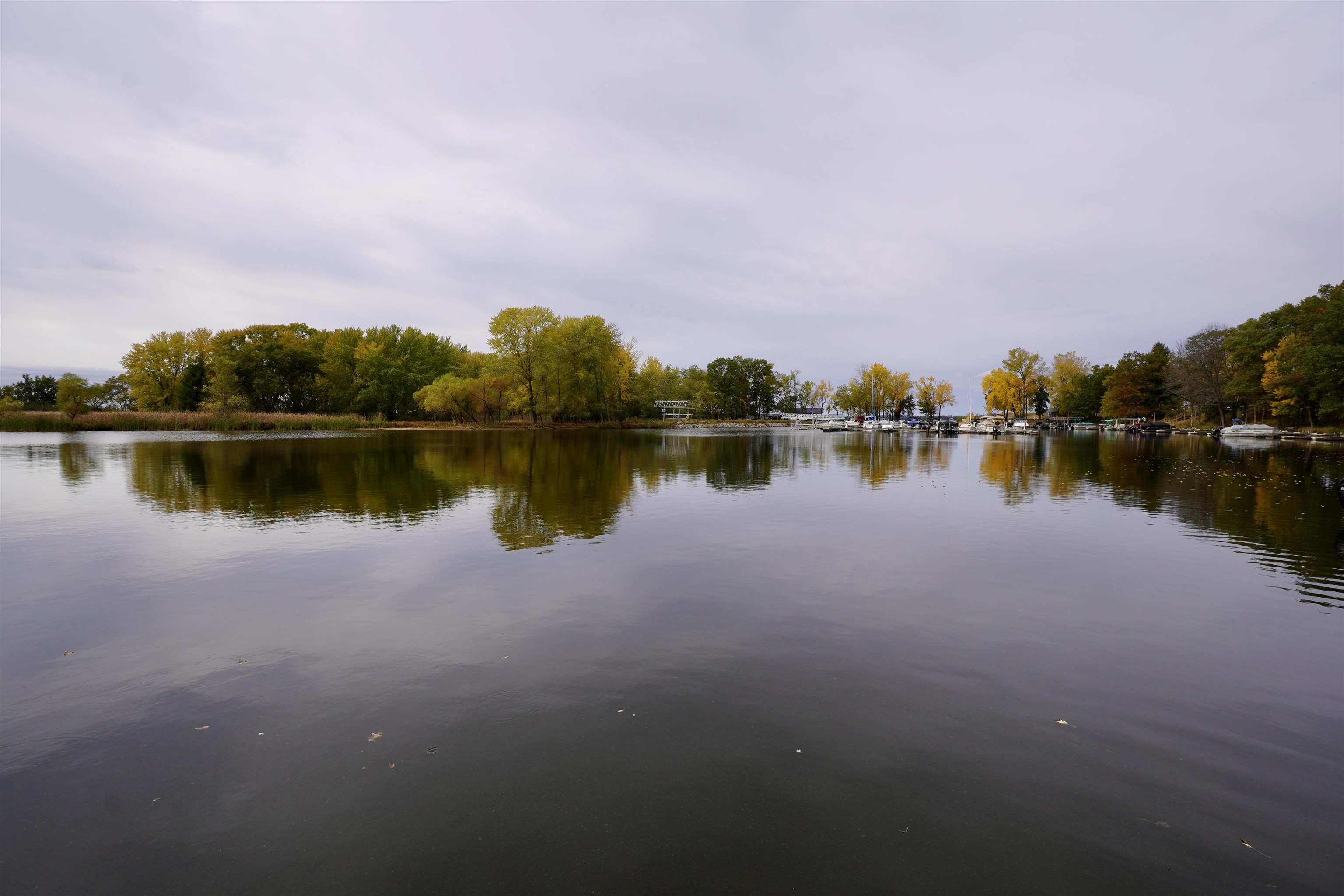 Calm lake reflecting autumn trees, with docks and boats along the distant shore.