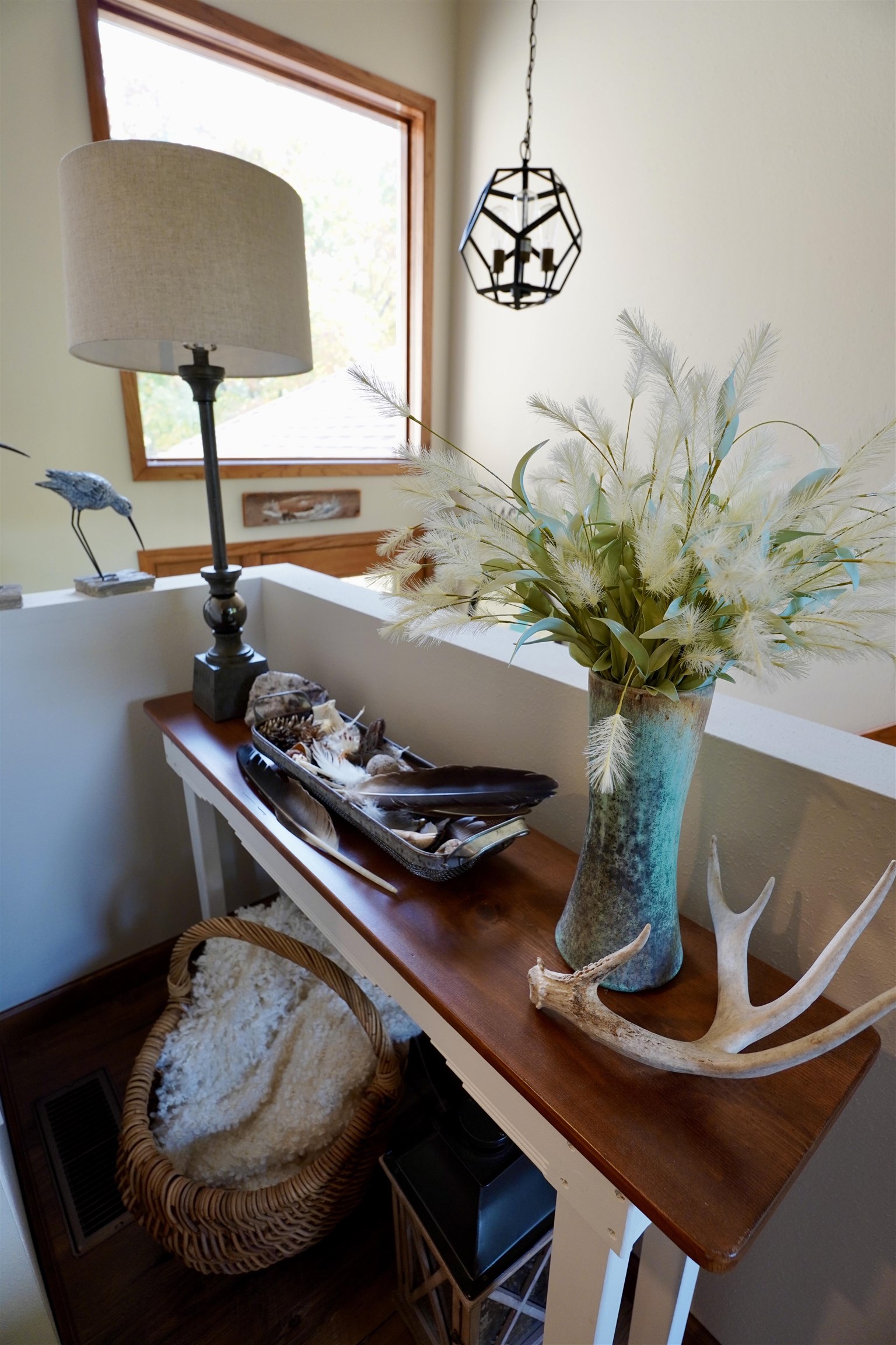 Interior landing with a wooden console table, turquoise vase, lamp, and antlers beside a large window.
