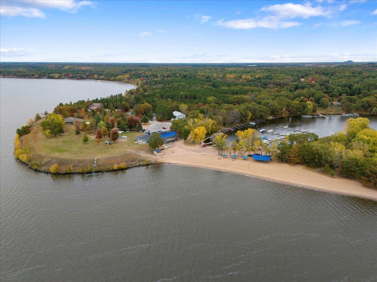 Aerial view of a wooded lakeside property, marina docks, sandy beach, and blue-roofed building.