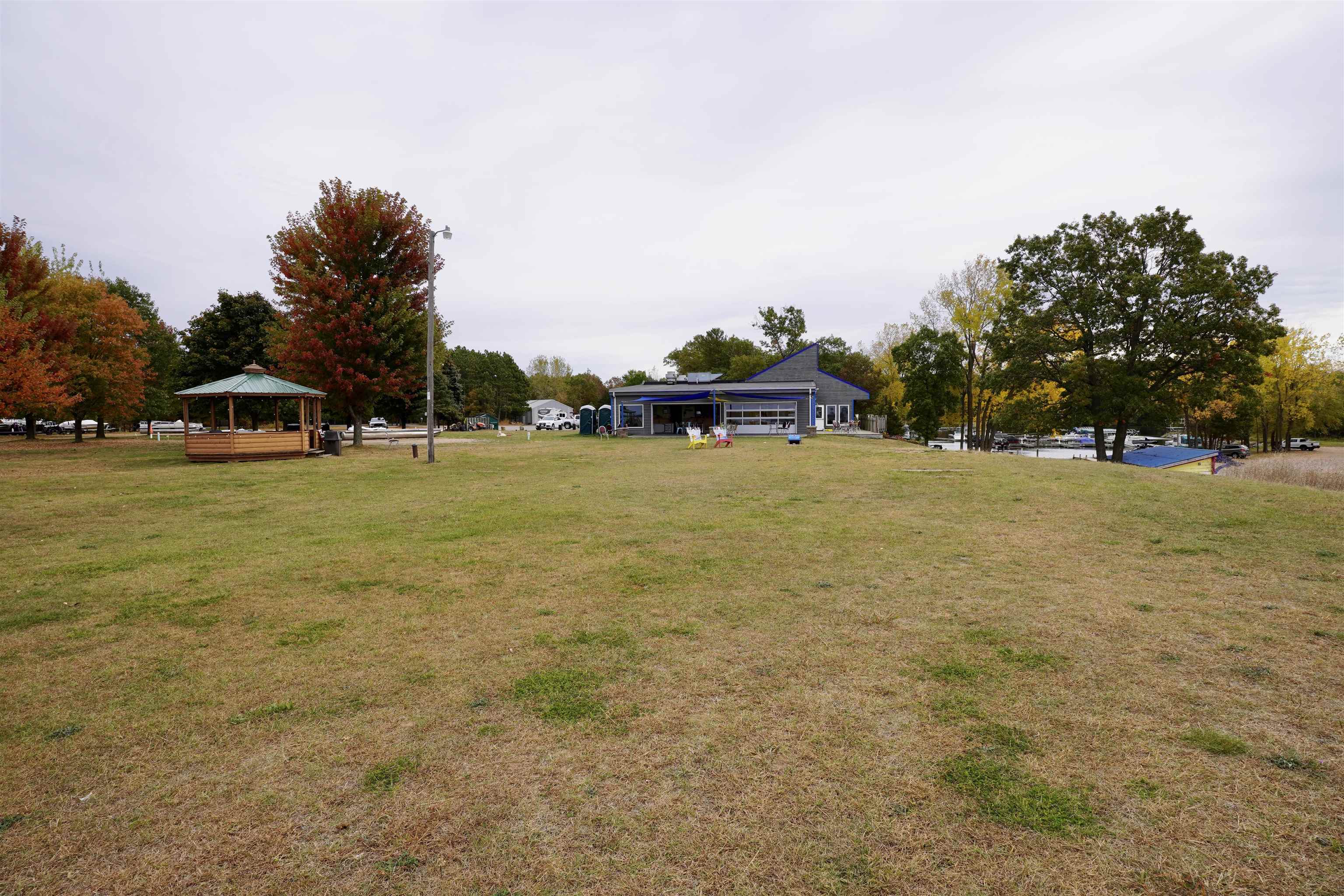 Lakeside lawn with a wooden gazebo, blue-trimmed building, boats, and autumn trees.